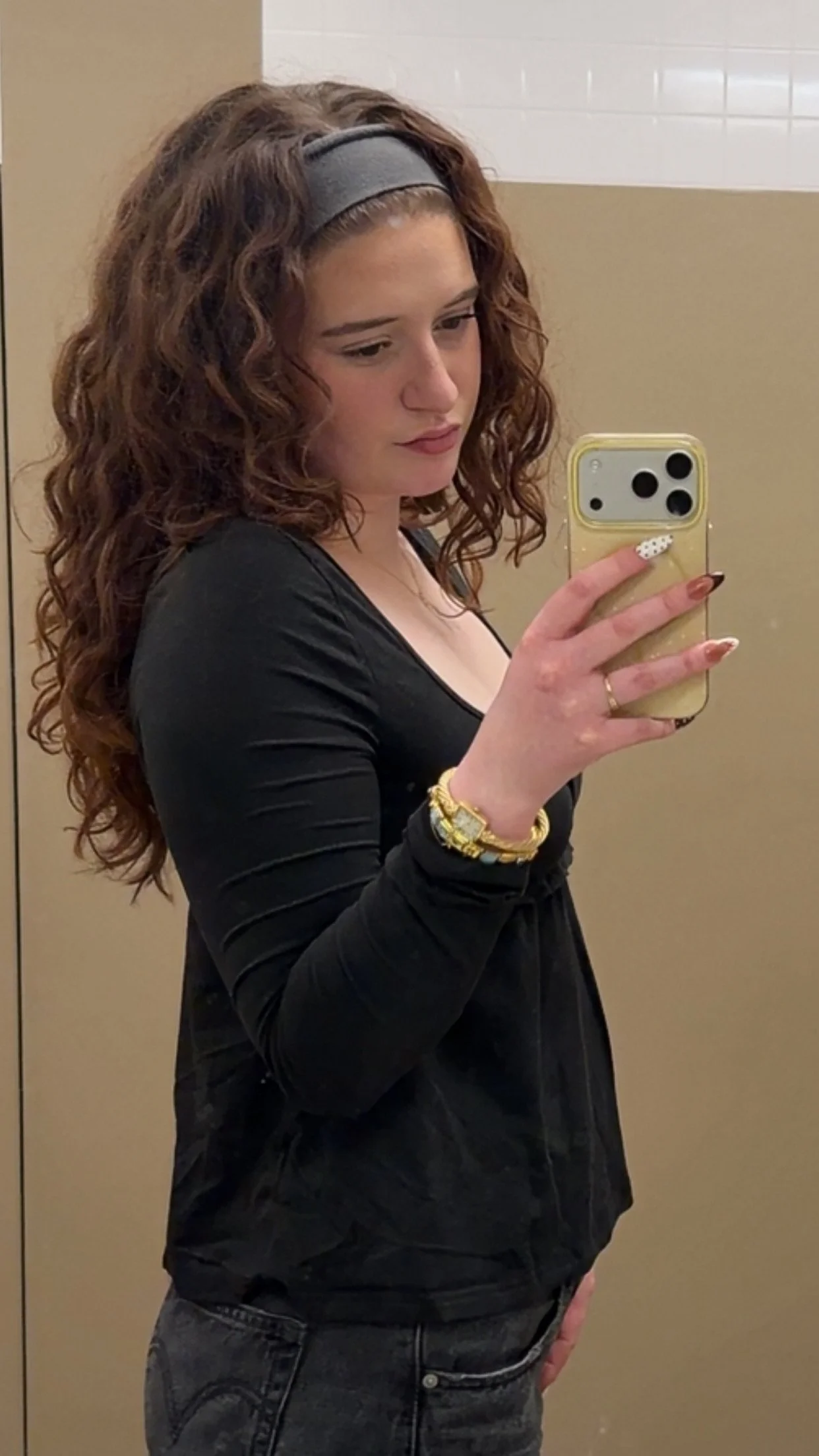 A young woman taking a mirror selfie in a restroom. She has curly brown hair with a headband, wearing a black top, gold watch, and bracelets.