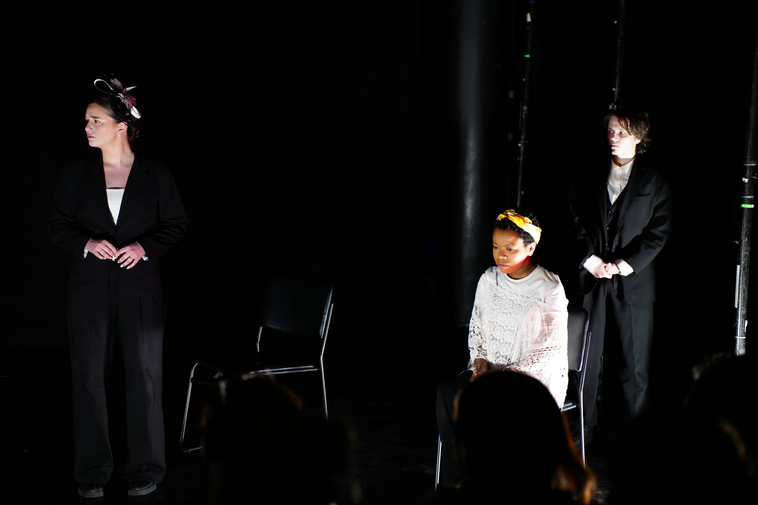 Three women on stage, two standing and one sitting, with a dark background and stage lighting.