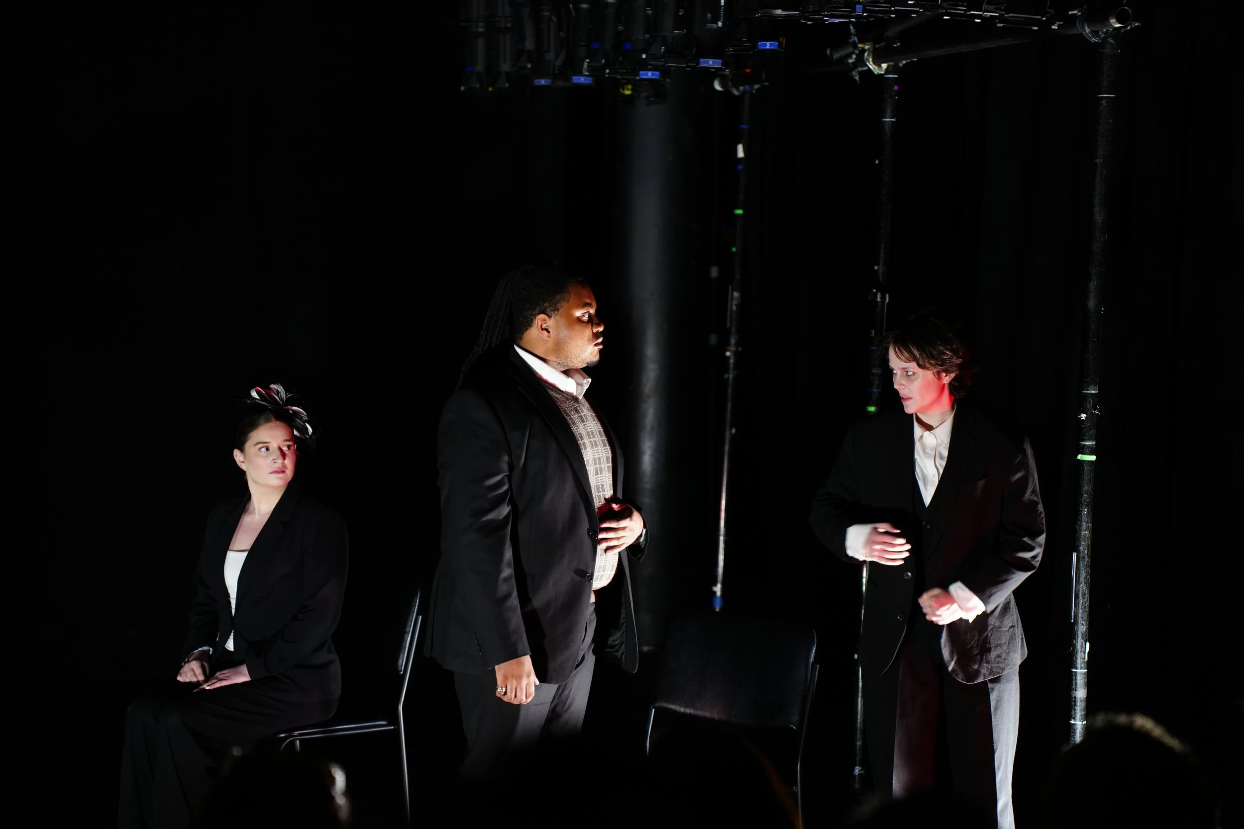 Scene with three people dressed in black suits, two women and one man, standing and sitting in a dark environment with stage lighting and equipment visible overhead.