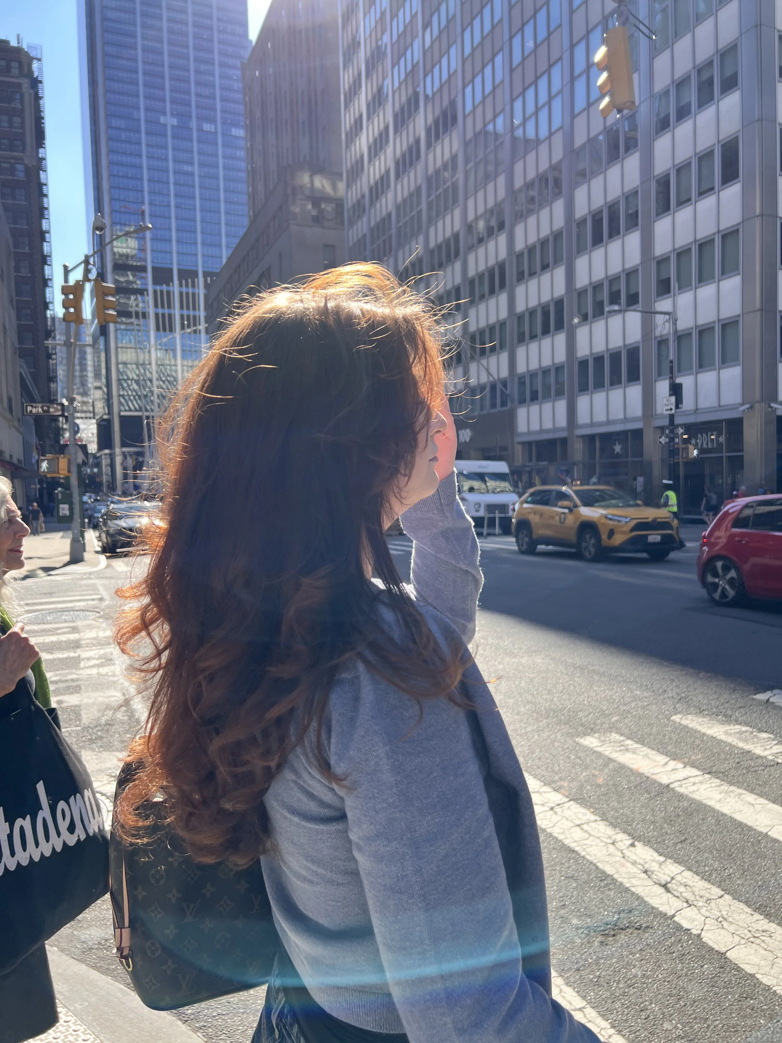 A person with long wavy brown hair wearing a gray jacket and carrying a Louis Vuitton backpack, standing on a city street with tall glass buildings and cars passing by.