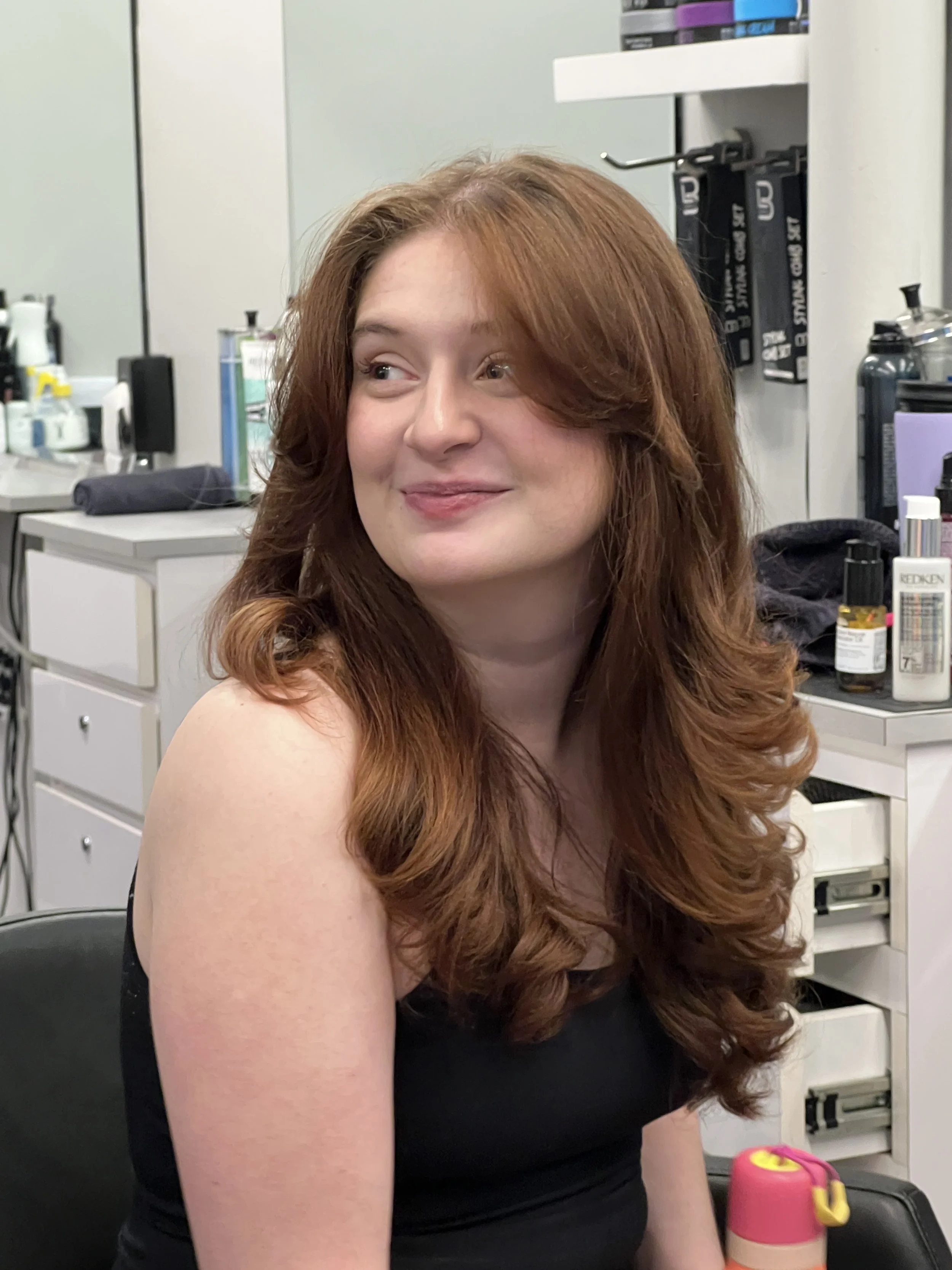 A woman with long red hair sitting in a room that looks like a medical or professional office. Shelves with various supplies and equipment are visible in the background.
