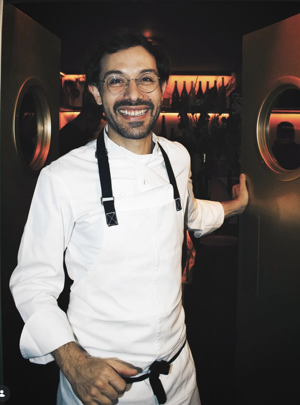 Chef sonriendo en la cocina, vestimenta blanca y con gafas, en un ambiente con iluminación cálida y decoración moderna.