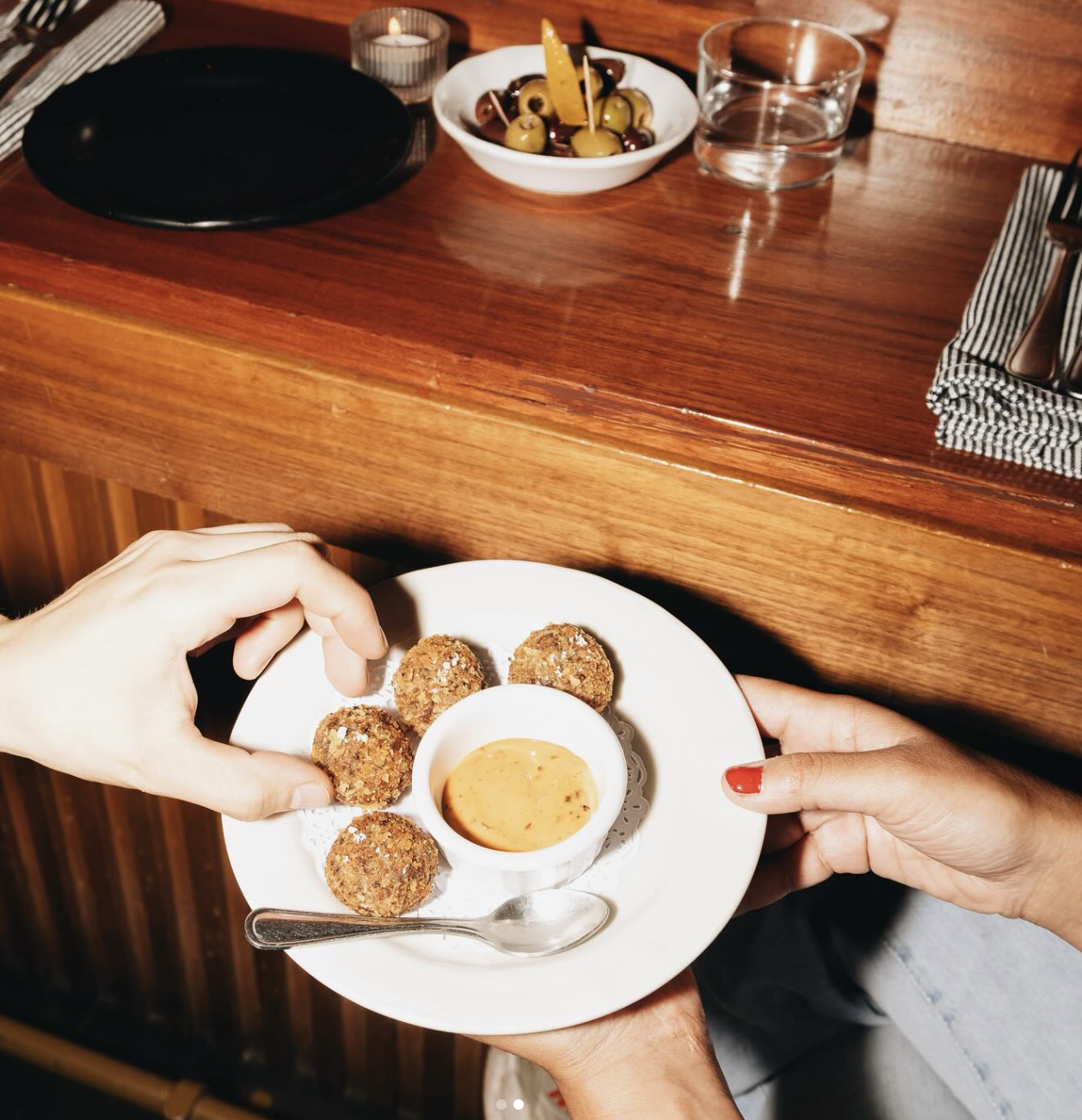 Manos sosteniendo un plato con cinco croquetas y una vasija con salsa en el centro, en un restaurante de madera. Platillo a base de hongos.
