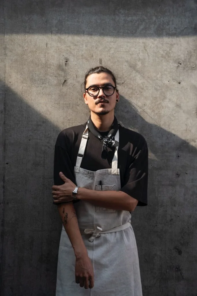 Joven con barba, gafas y cabello enrollado, vistiendo un delantal y camisa negra, de pie frente a un muro de concreto, con luz que crea una sombra diagonal. Ricardo Verdejo.