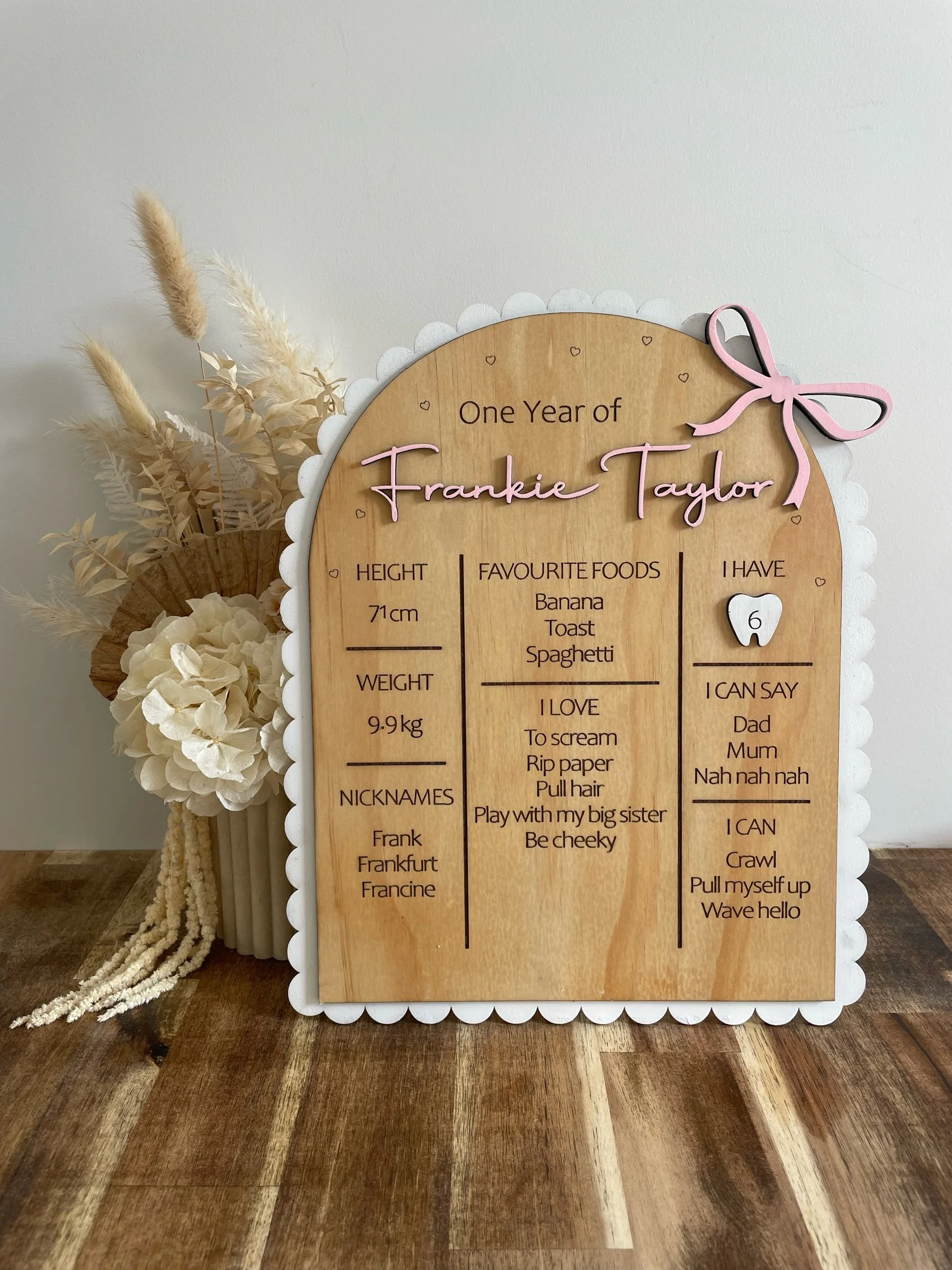A wooden milestone board decorated with pink and white accents, celebrating a child's first year. It has sections for height, favorite foods, and things the child can say, along with labels for nicknames and other personal details. There are dried flowers and beige decorations arranged beside it.