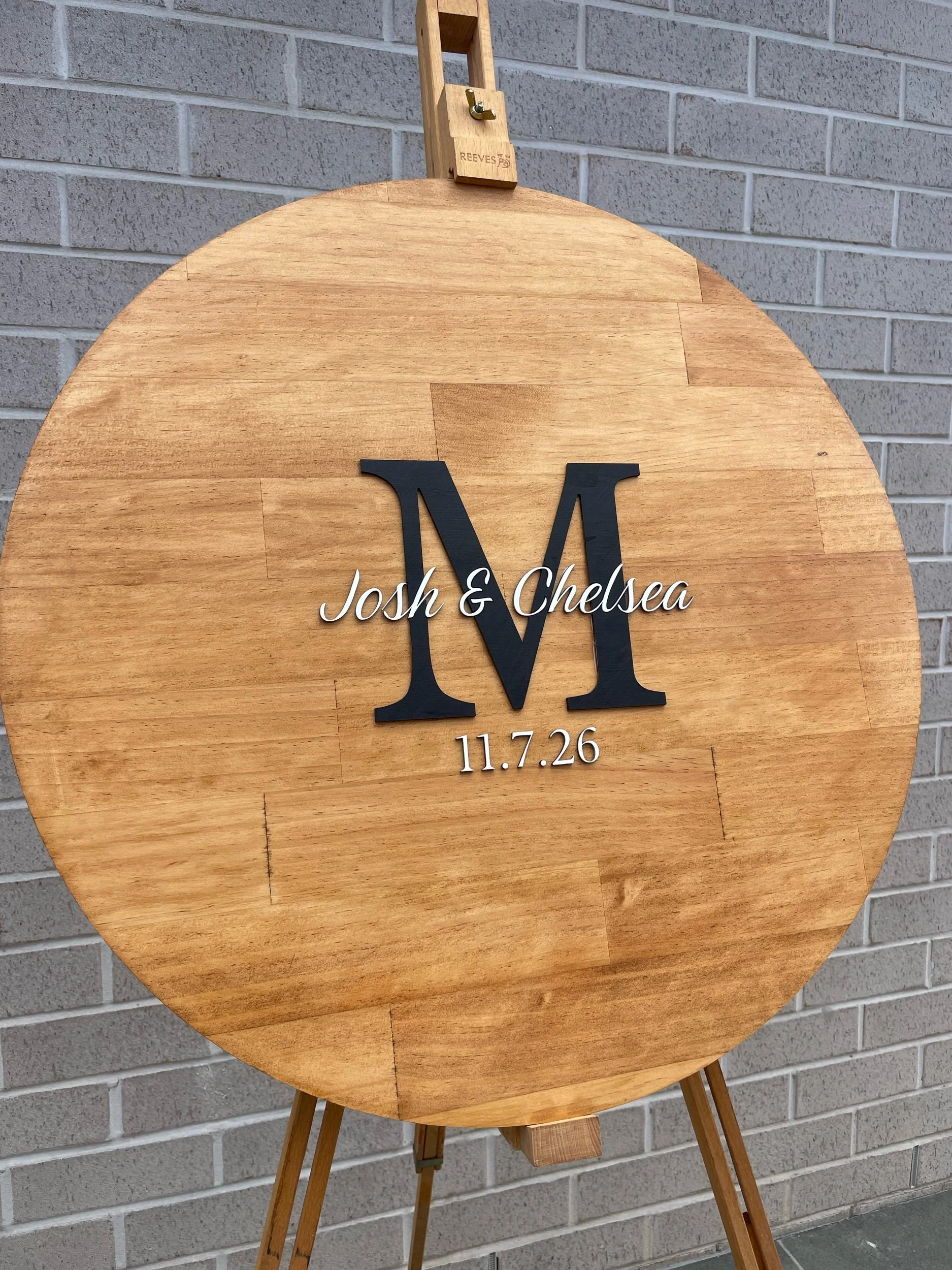 Wooden wedding sign with black and white lettering displaying the initials 'M' and 'W', the names 'Josh & Chelsea', and the date '11.7.26', against a brick wall backdrop.