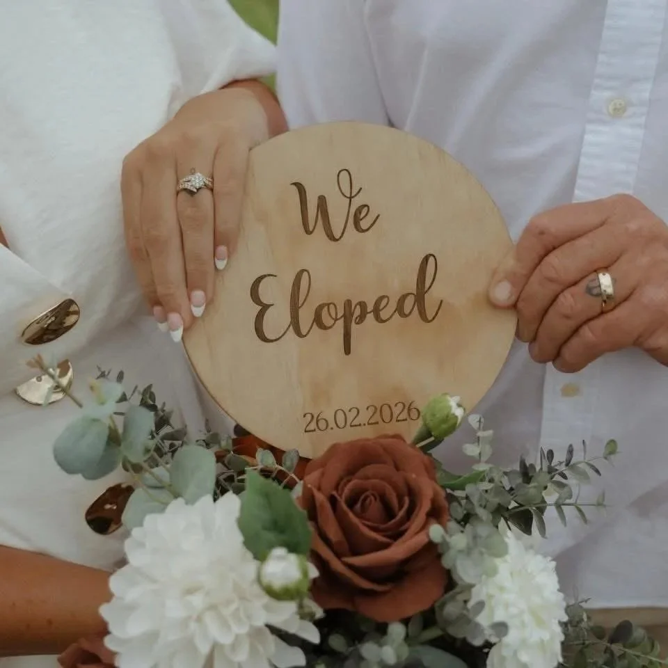 Two people holding a wooden sign that says "We Eloped" with a date below, surrounded by flowers.