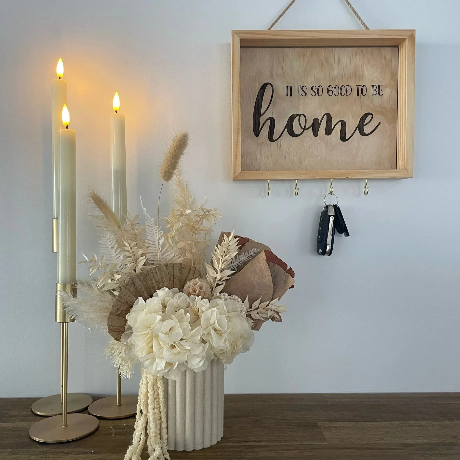 Decorative display including three lit candles, beige pampas grass, dried flowers, and a floral arrangement on a dark wood surface, next to a wall-mounted wooden sign with the phrase 'It is so good to be home'.