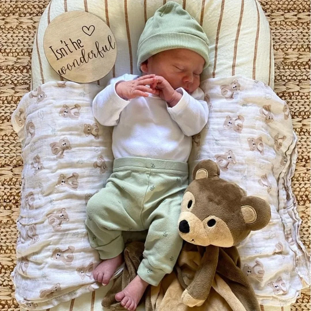 A sleeping newborn baby wearing a green beanie, white shirt, and mint green pants, lying on a pillow with bear print. The baby is holding hands together and is accompanied by a brown bear plush toy. A wooden round sign with the phrase 'Isn't the wonderful' is beside the baby.