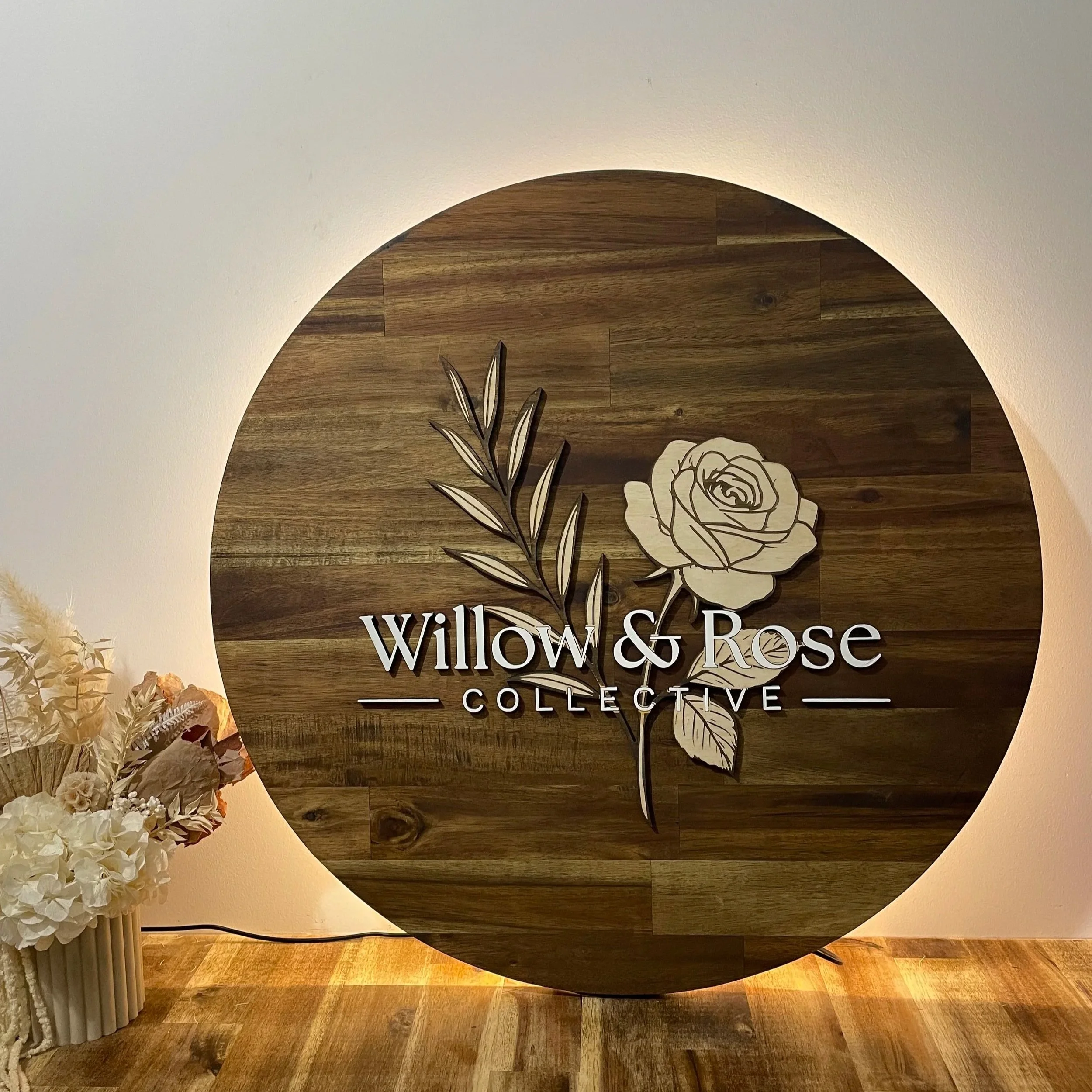 Wooden round sign with floral illustration and the text 'Willow & Rose Collective' on a wall with subdued lighting.