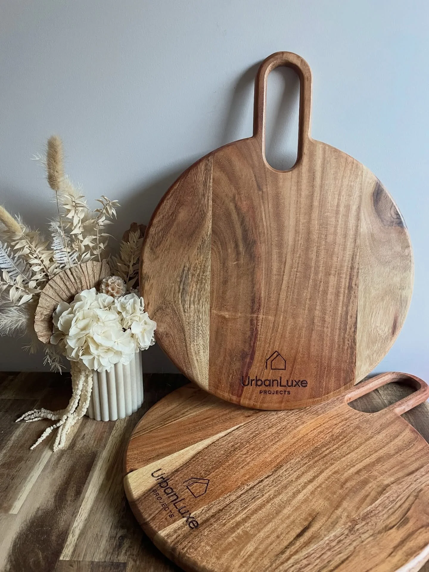 Round wooden cutting boards leaning against a white wall, with one featuring the logo "UrbanLuxe Projects". There is a white vase with white flowers and natural dried plants beside the boards.