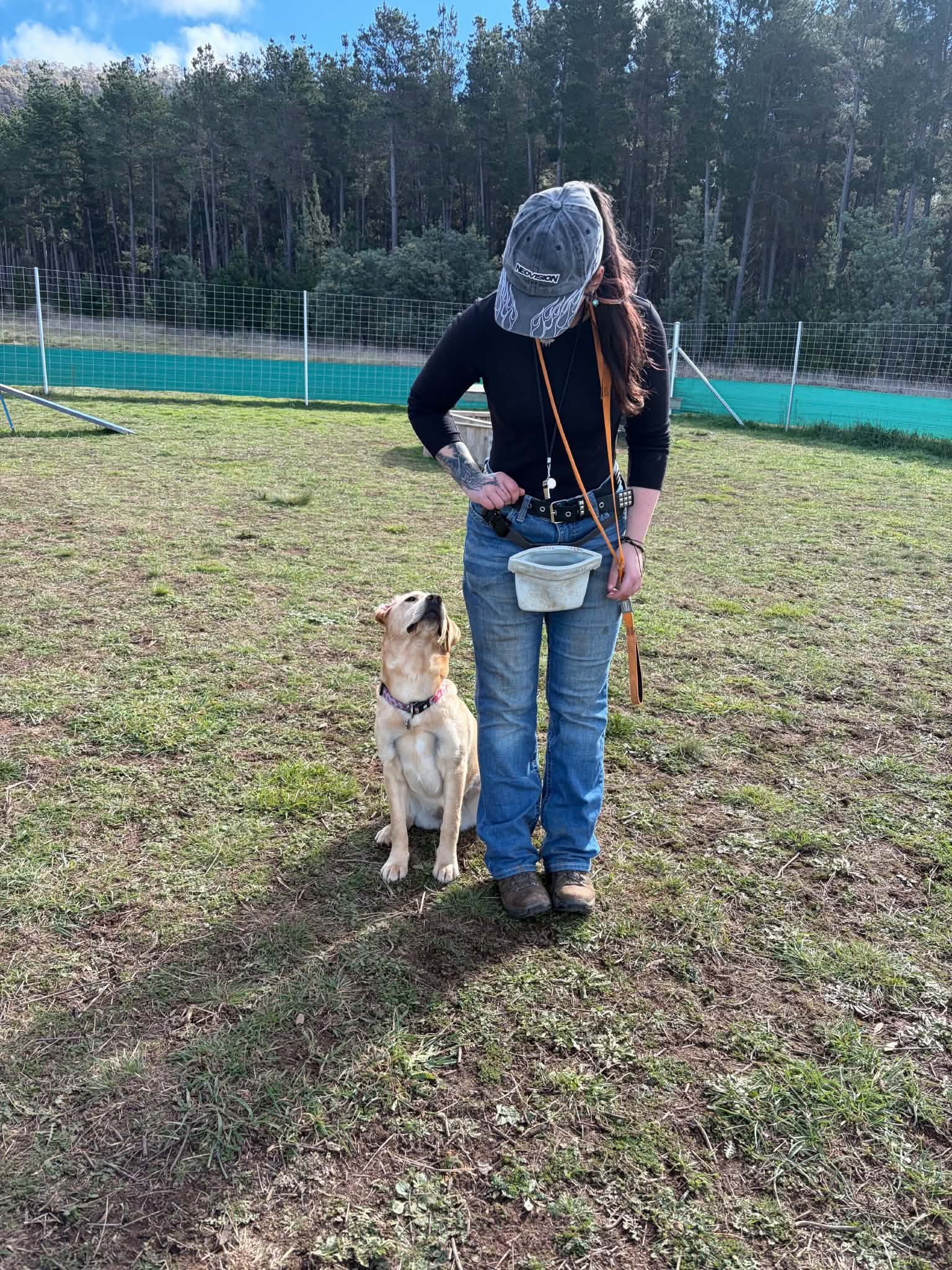 A person with long dark hair wearing a gray cap, black long-sleeve shirt, blue jeans, and black boots, standing outdoors with a yellow Labrador Retriever puppy, on a grassy field with a chain-link fence and trees in the background.
