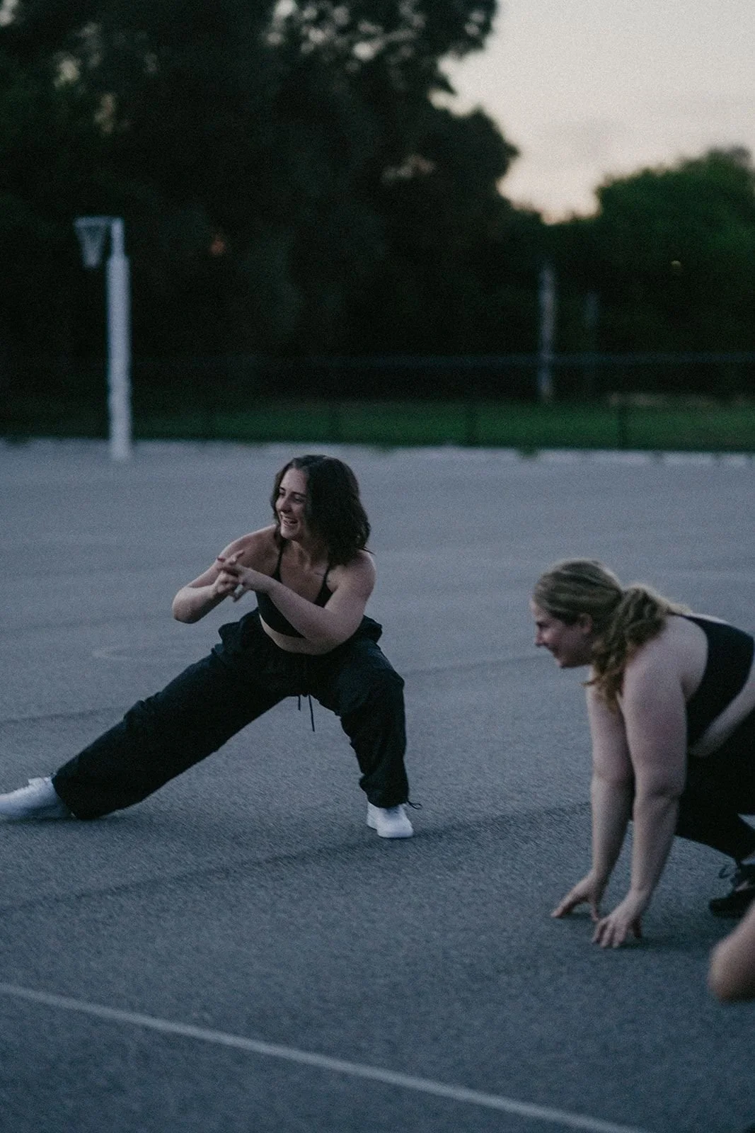 Two women outdoors on a basketball court during dusk, smiling and stretching or warming up.