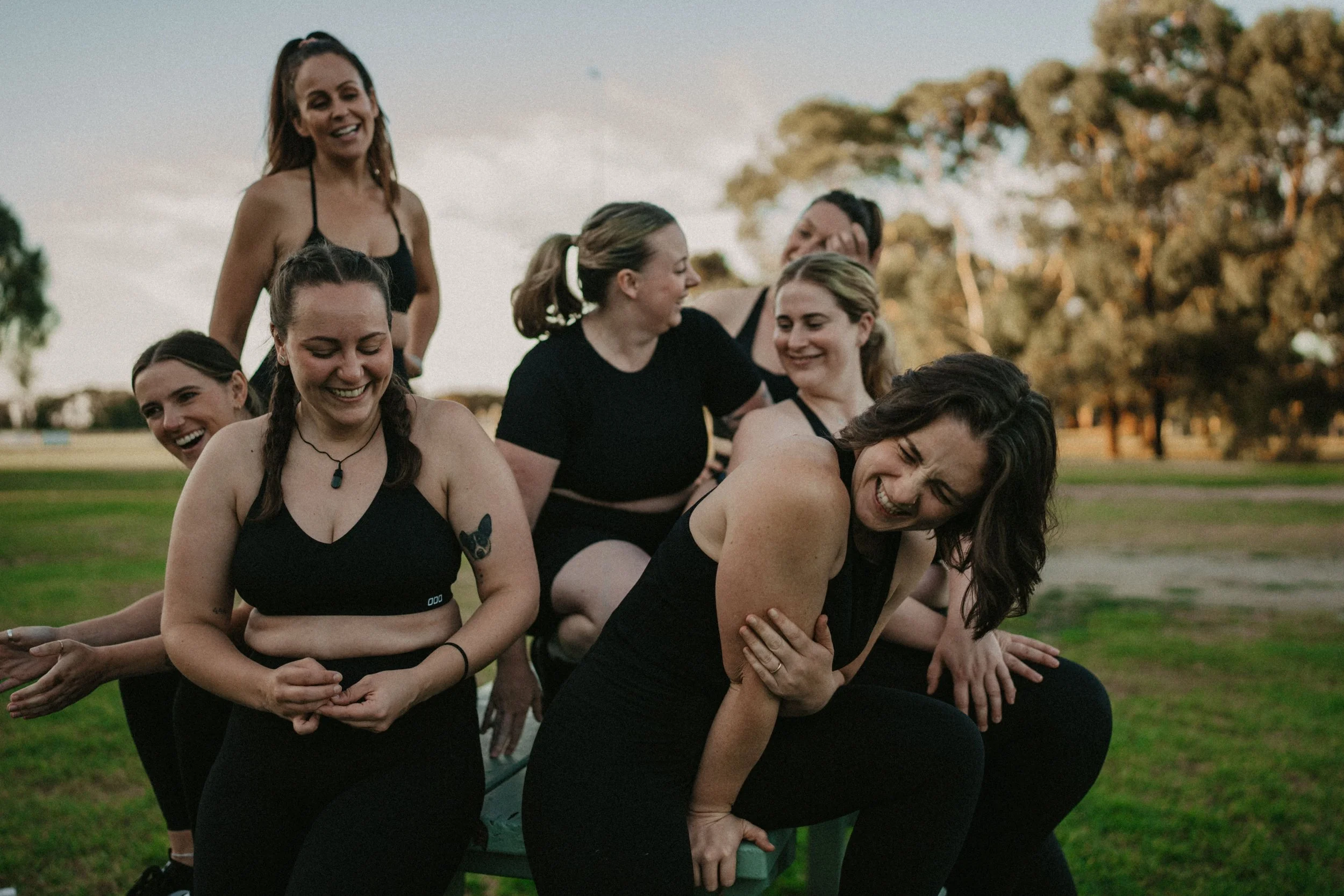 A group of women practicing a yoga pose outdoors in a park during the daytime, smiling and interacting with each other.
