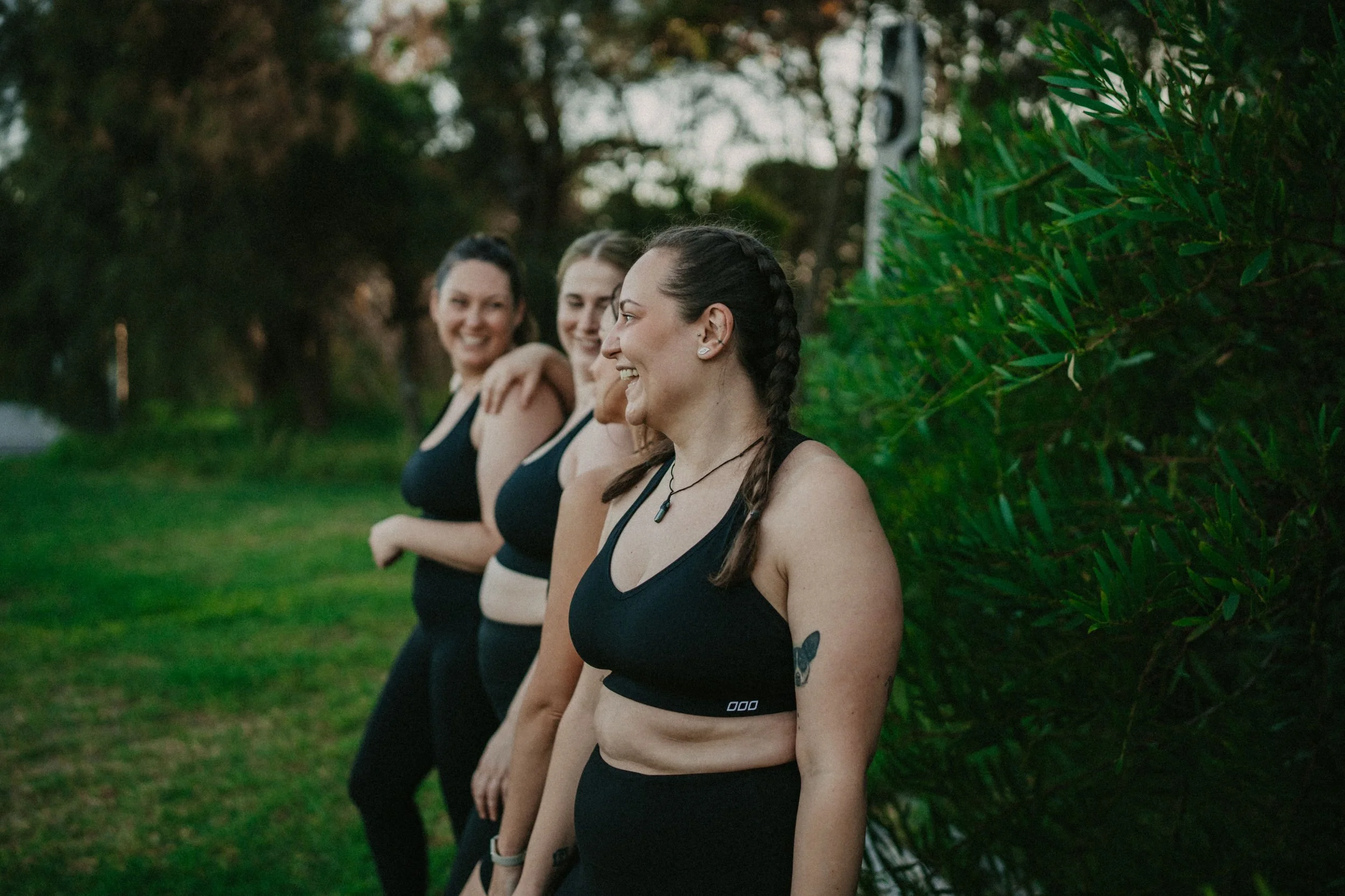 Four women standing outdoors in athletic attire, smiling, with a background of trees and bushes.