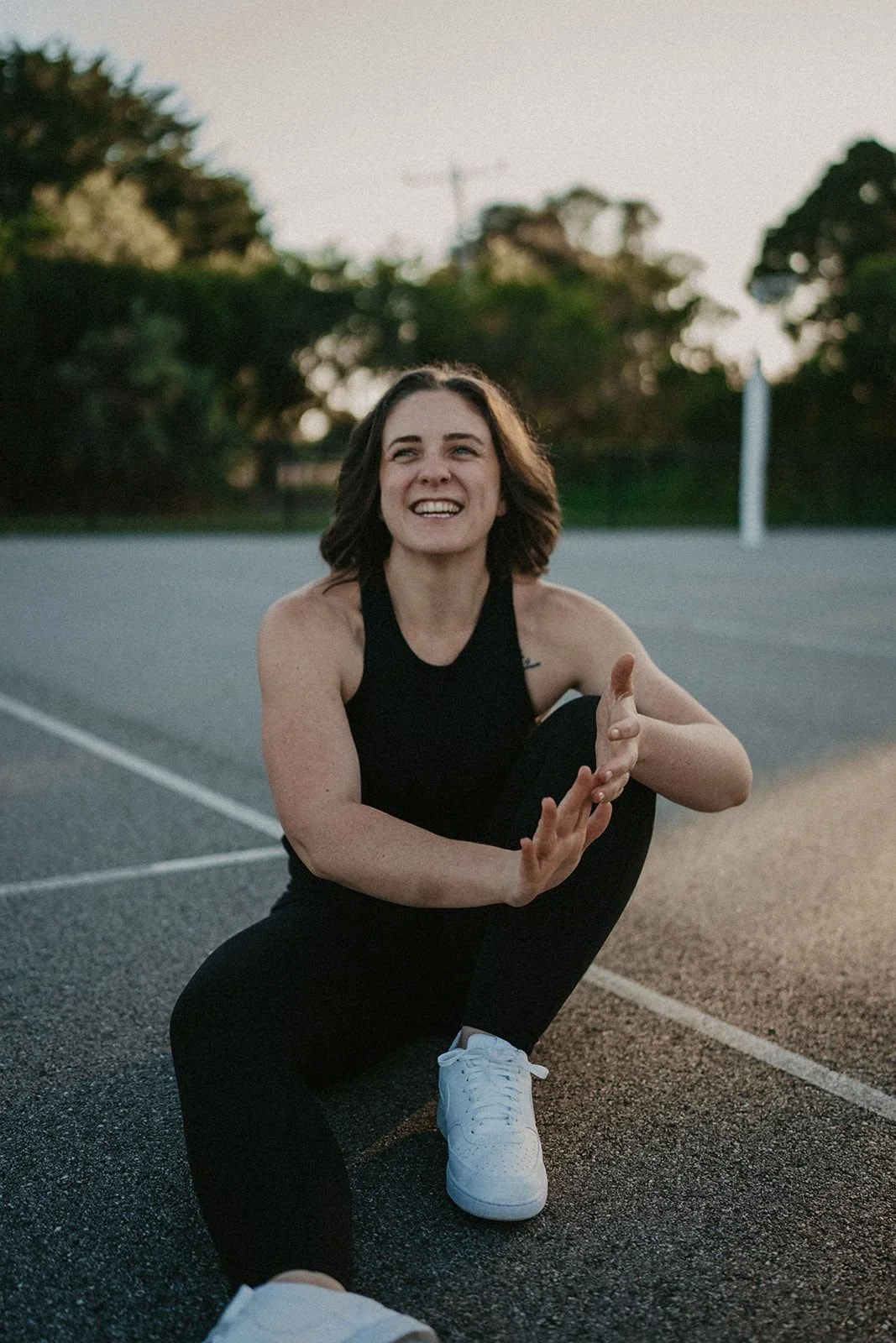 A woman in athletic clothing, smiling and stretching on an outdoor basketball court during sunset.