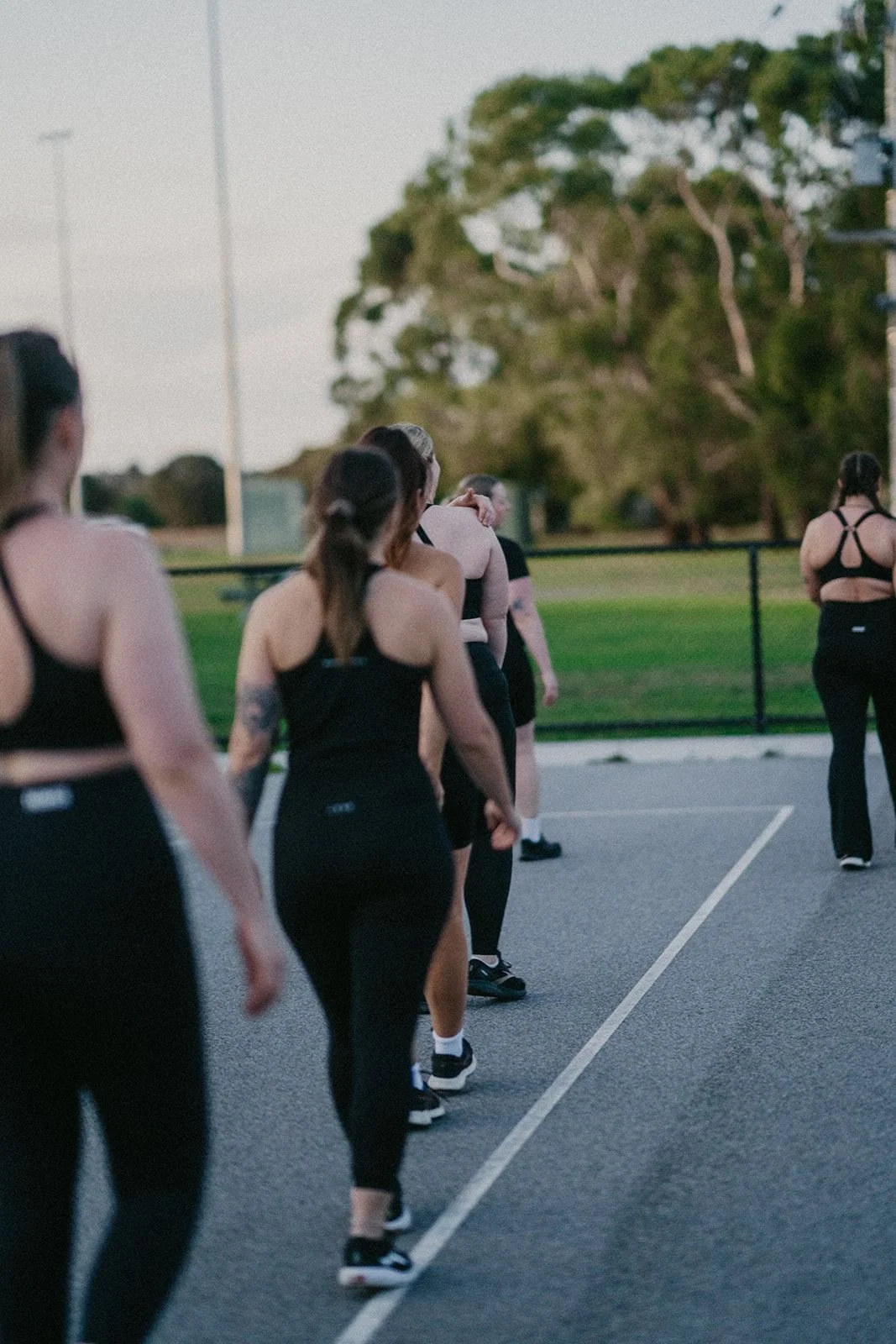 Group of women participating in an outdoor fitness or dance class on a basketball court during the evening, with trees and a fence in the background.