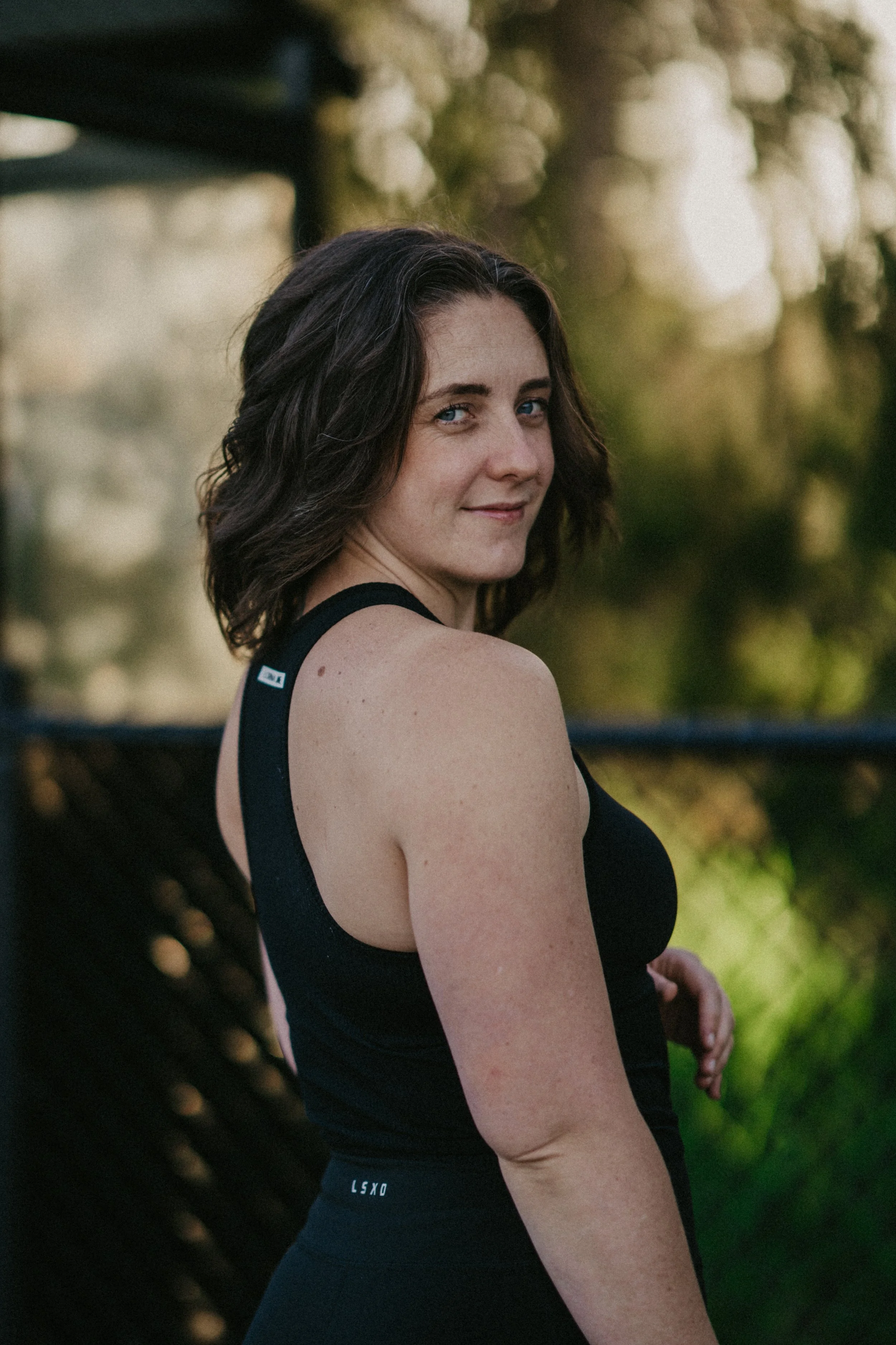 A woman with wavy dark brown hair and blue eyes, wearing a black athletic tank top, standing outdoors near a wooden structure, with a blurred background of trees and sunlight.