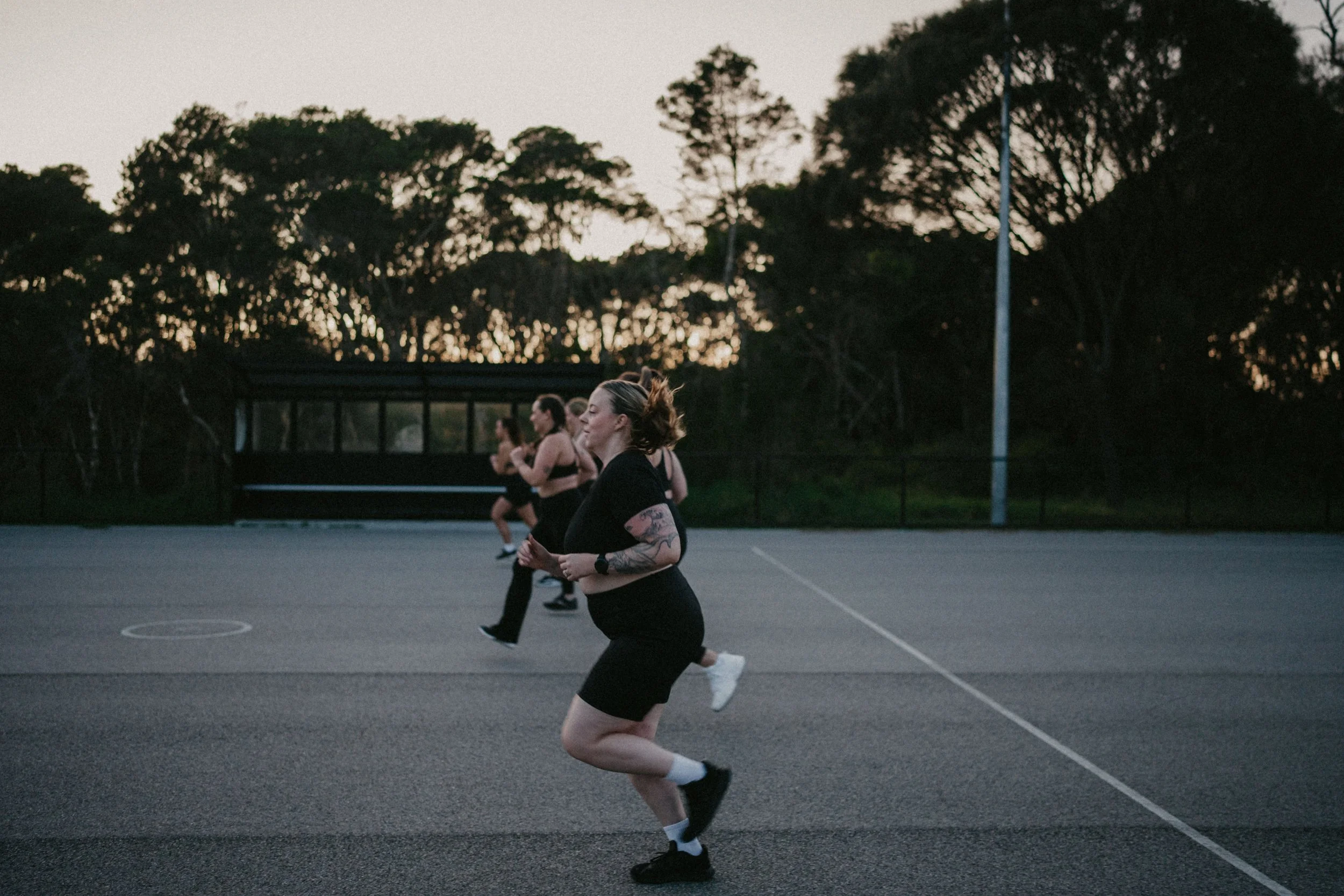 Women running on an outdoor basketball court during dusk, with trees and light poles in the background.