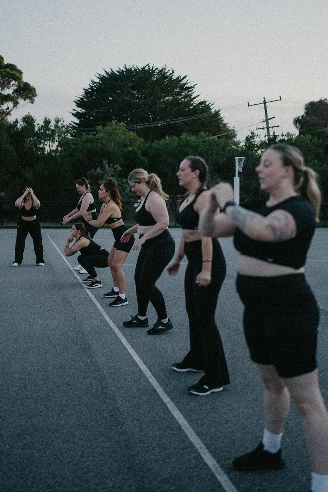Group of women exercising outdoors on a basketball court during dusk, in athletic wear, with trees and utility poles in the background.