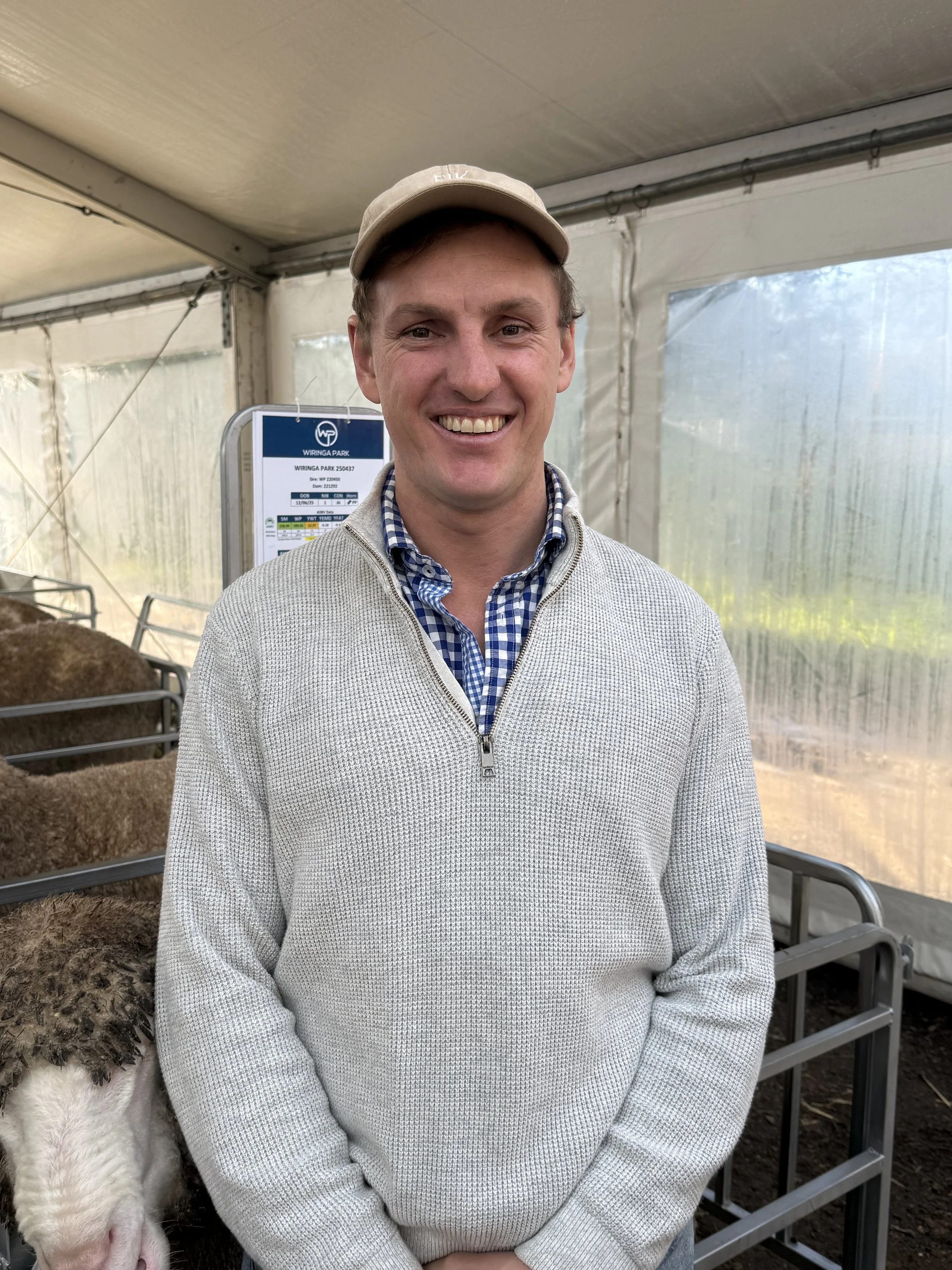Wiringa Team Photo. A smiling man in a gray sweater and plaid shirt standing inside a tent or greenhouse, with some pigs in pens behind him.