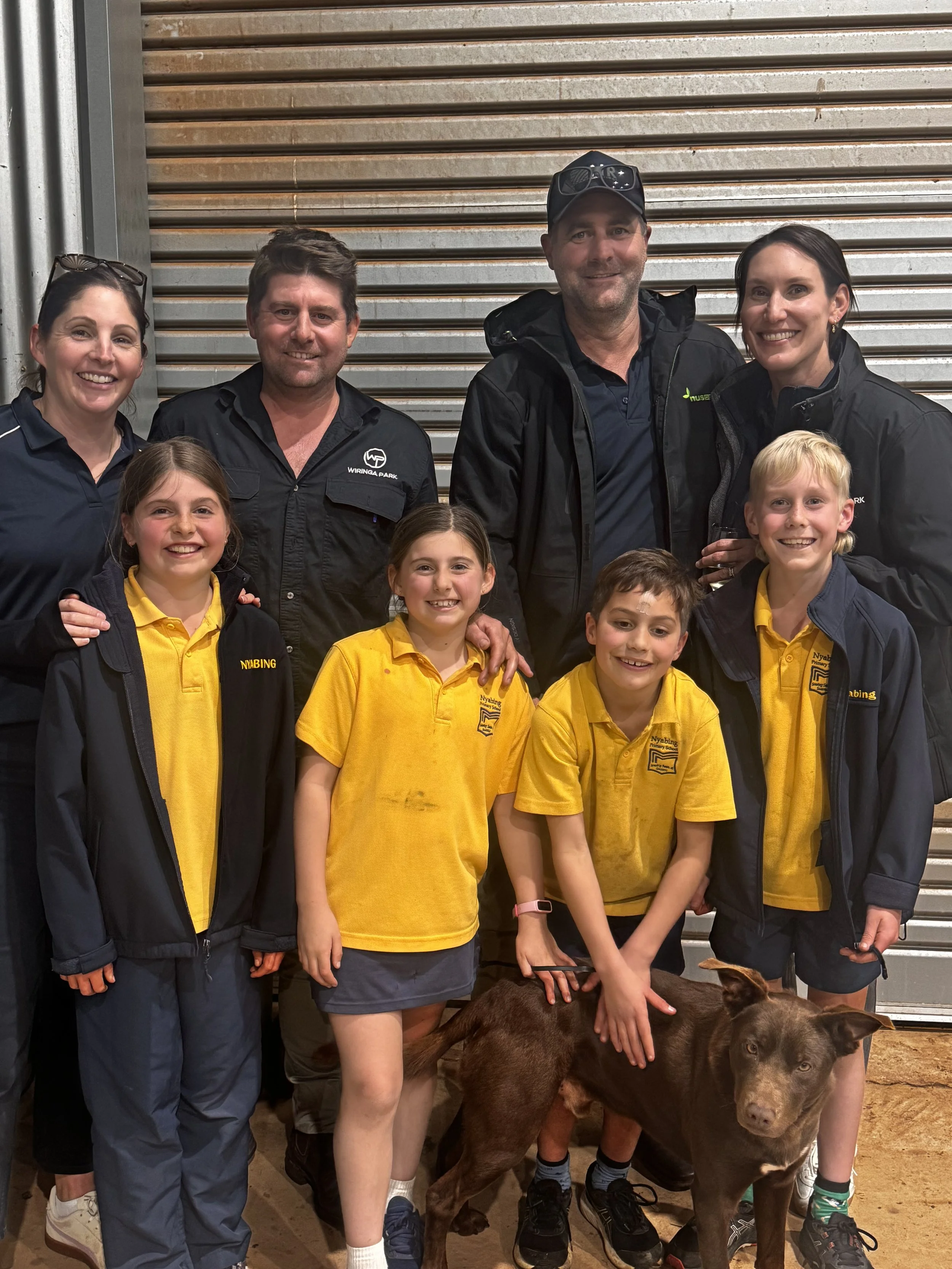 Wiringa Team, two families behind Wiringa Park. Group of seven people, including children and adults, posing indoors with a brown dog in front of a metal roller door.