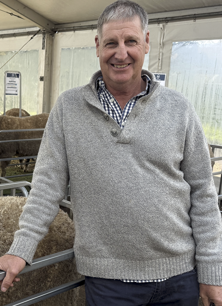 Wiringa Team Photo. A smiling man in a gray sweater and checkered shirt standing inside a tent with sheep or goats behind a metal fence.