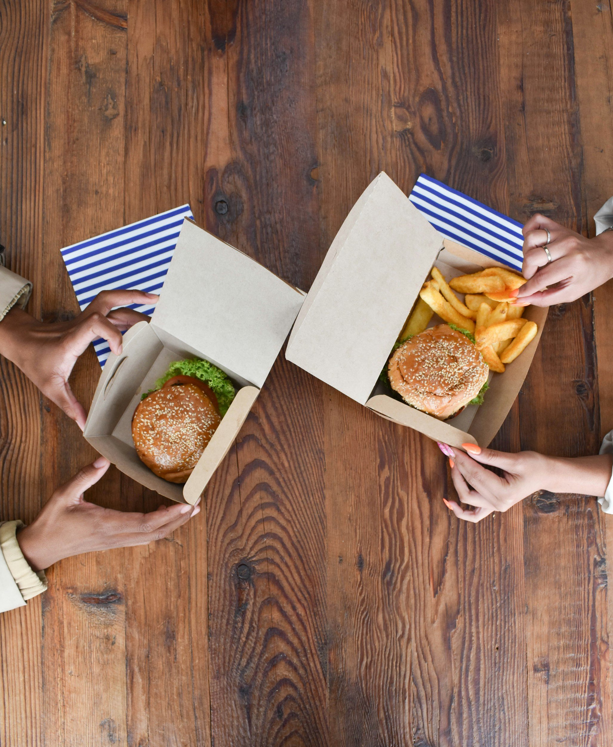 Top-down view of people holding and opening fast food takeout containers with burgers, fries, and lettuce on a wooden table.