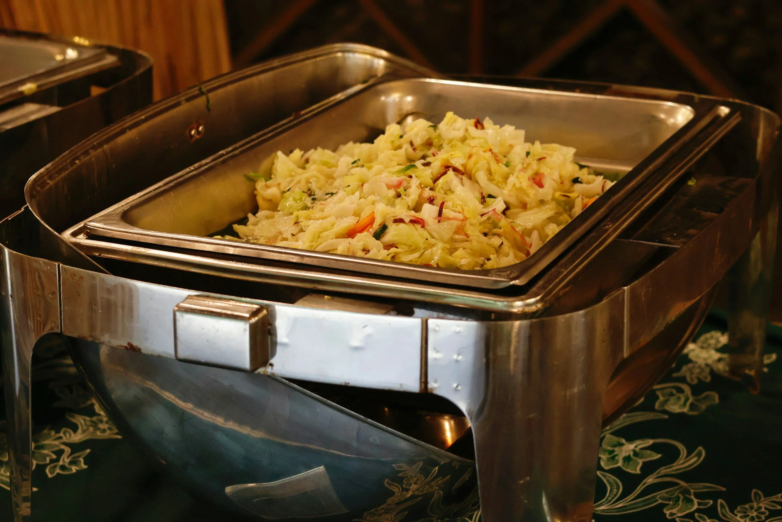 A stainless steel chafing dish filled with a mix of cooked vegetables, including cabbage, carrots, and possibly peppers, sitting on a decorative cloth with a dark background.