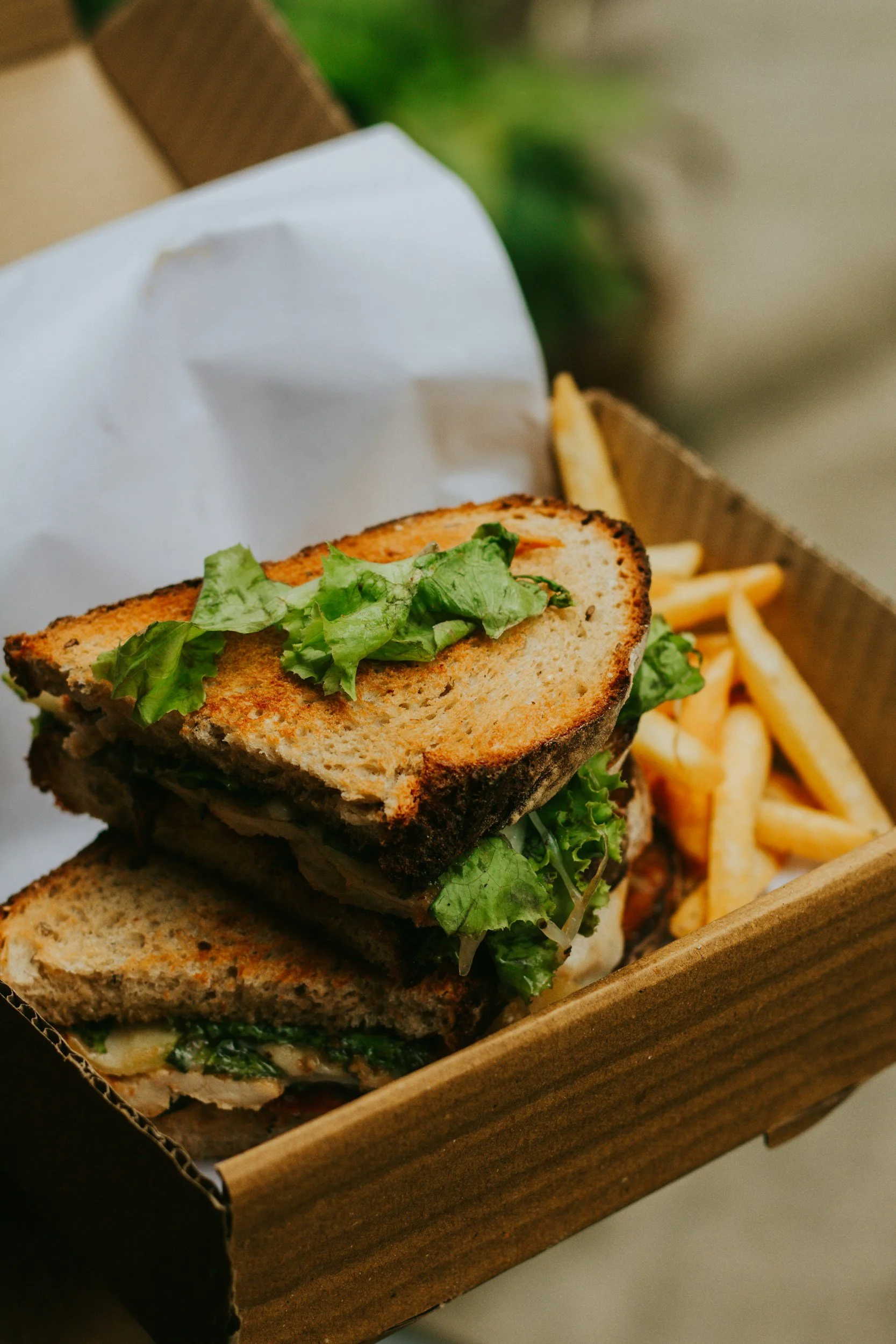 A close-up of a sandwich with grilled bread, lettuce, and other ingredients, placed in a wooden container with French fries in the background.