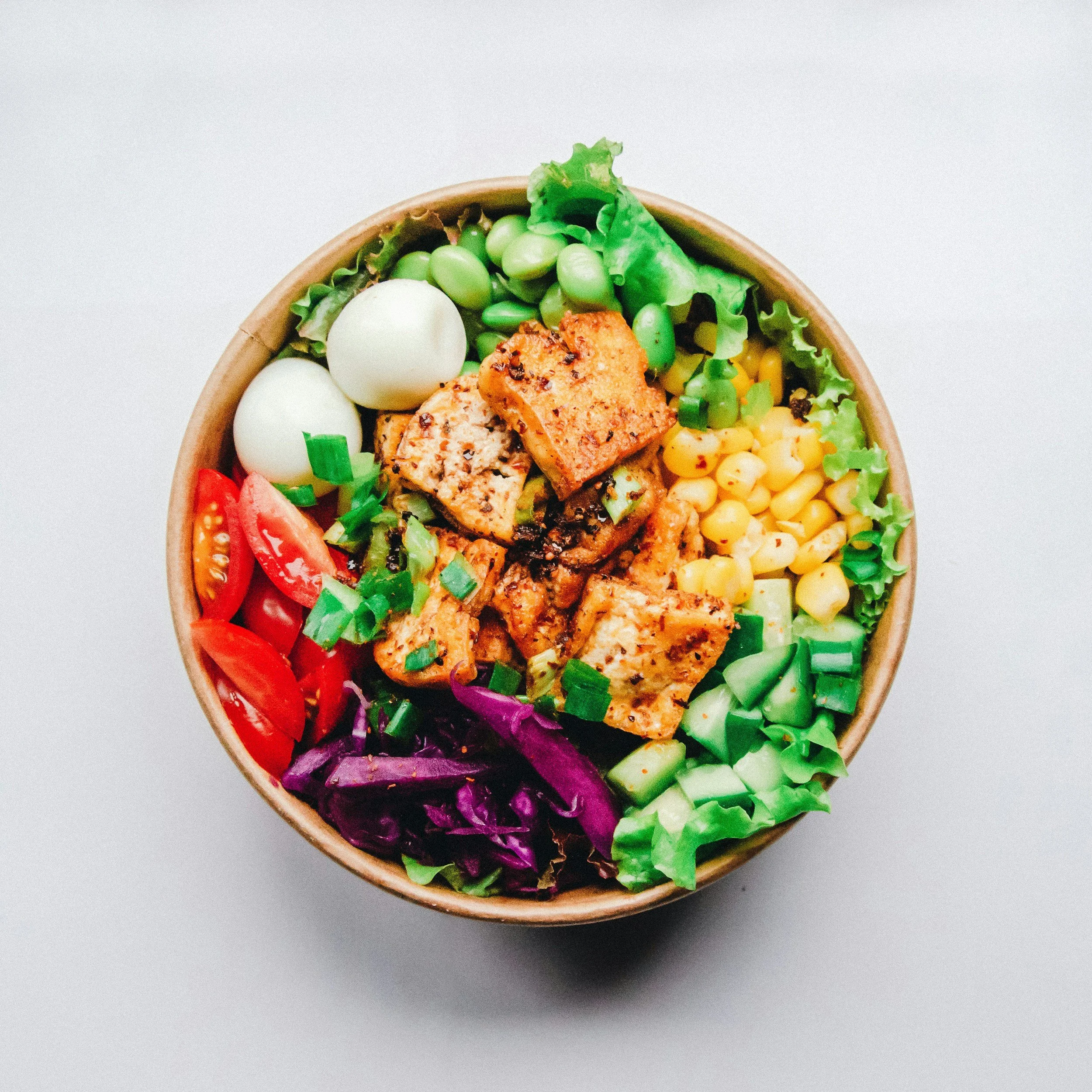 A bowl of salad with cherry tomatoes, boiled eggs, grilled chicken, corn, various greens, and chopped vegetables.