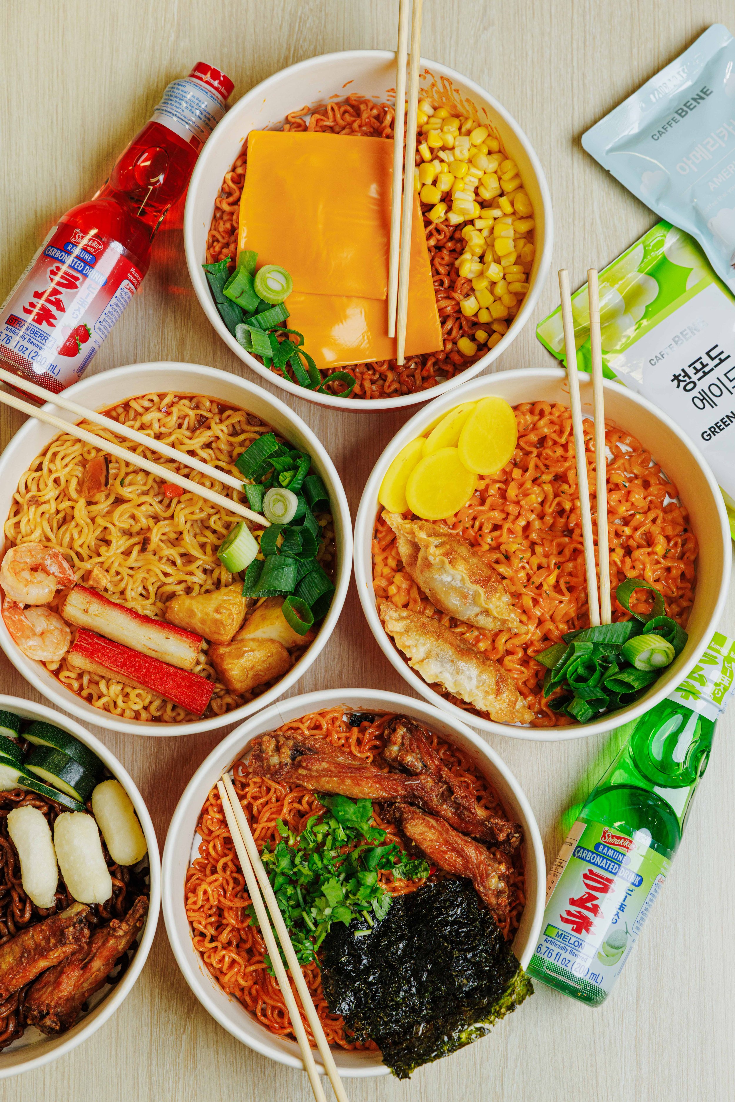 Various bowls of Korean instant noodles topped with ingredients such as cheese slices, corn, green onions, and fried items, with bottles of carbonated drinks on a wooden table.