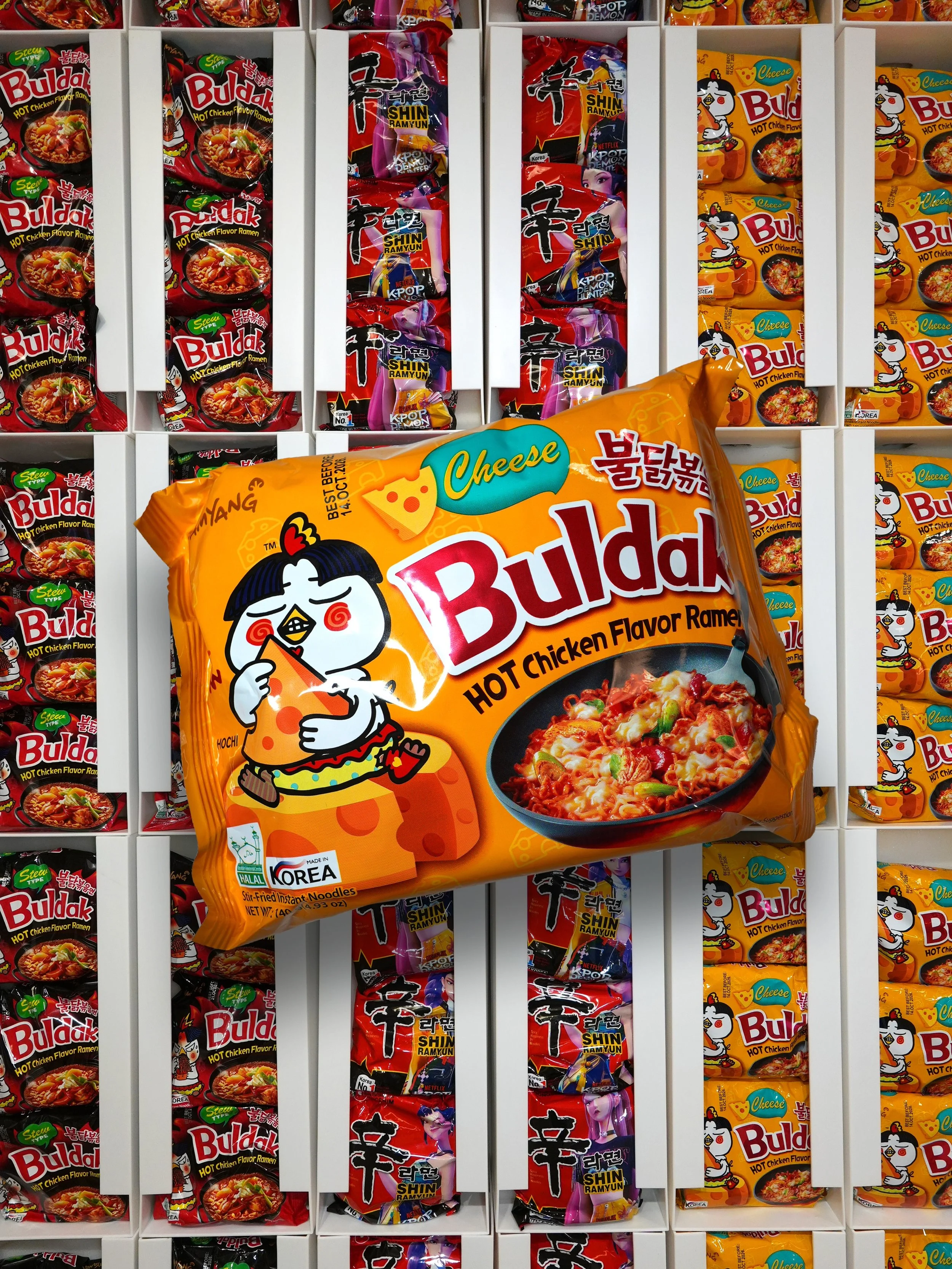 A shelf filled with various flavors of instant ramen packets, including Buldak spicy chicken flavor, Shin Ramyun, and Cheesy Buldak, organized in white cubbies.