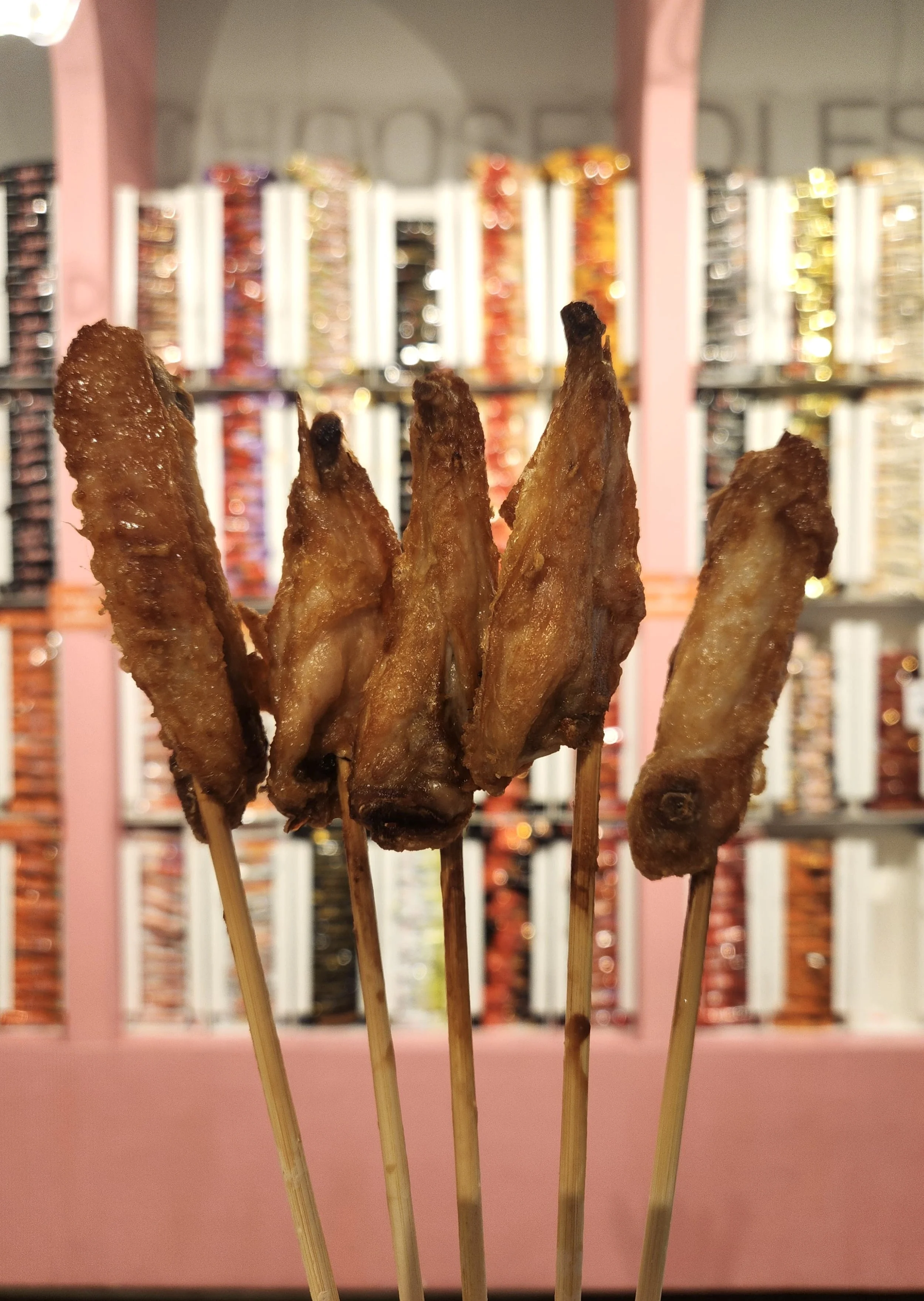Close-up of five grilled meat skewers in the foreground, with a blurred background of nail polish displays in a store or salon.