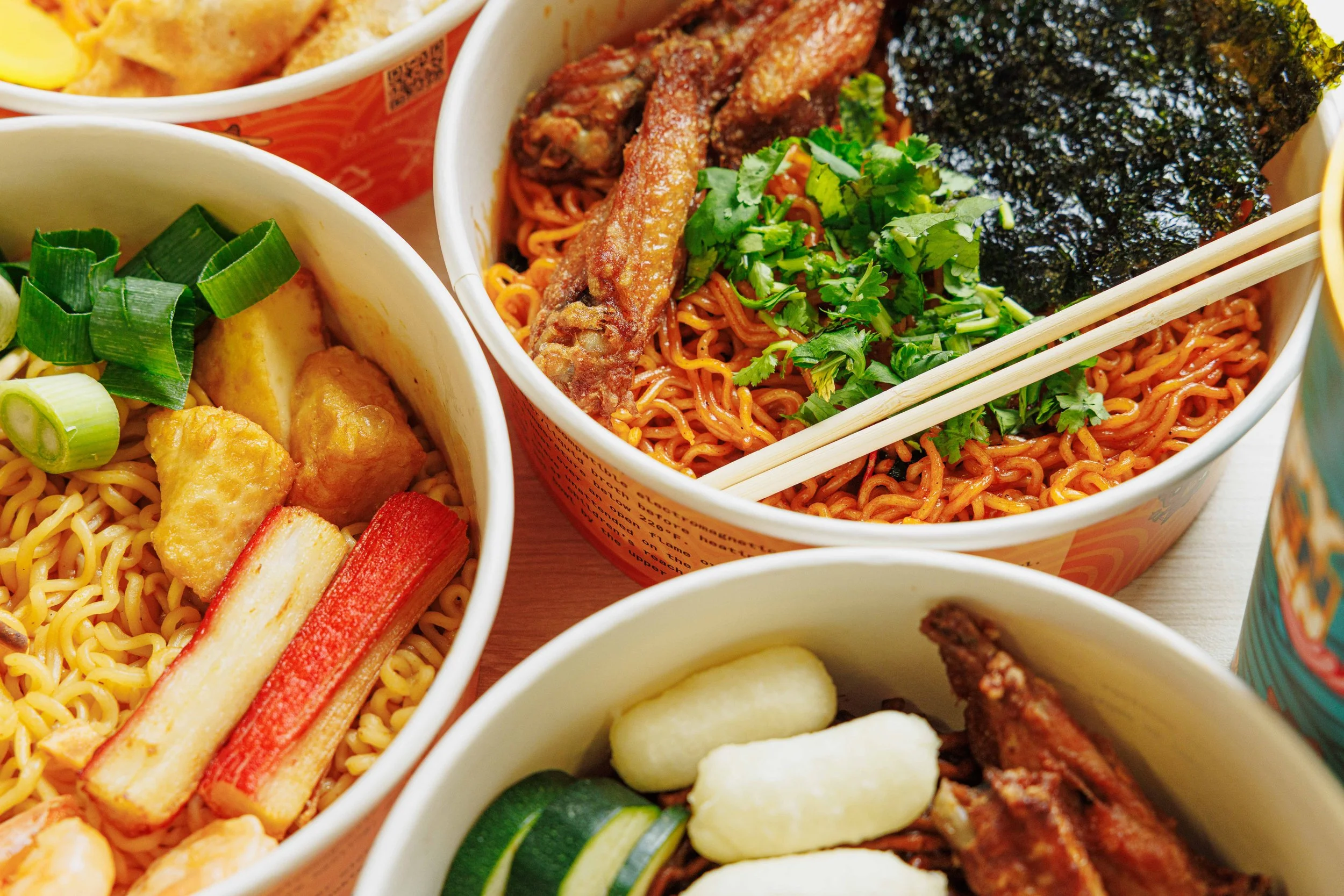 Close-up view of assorted Asian-style noodle bowls filled with ingredients like fried chicken, seaweed, green onions, cilantro, cucumber slices, and crab sticks.