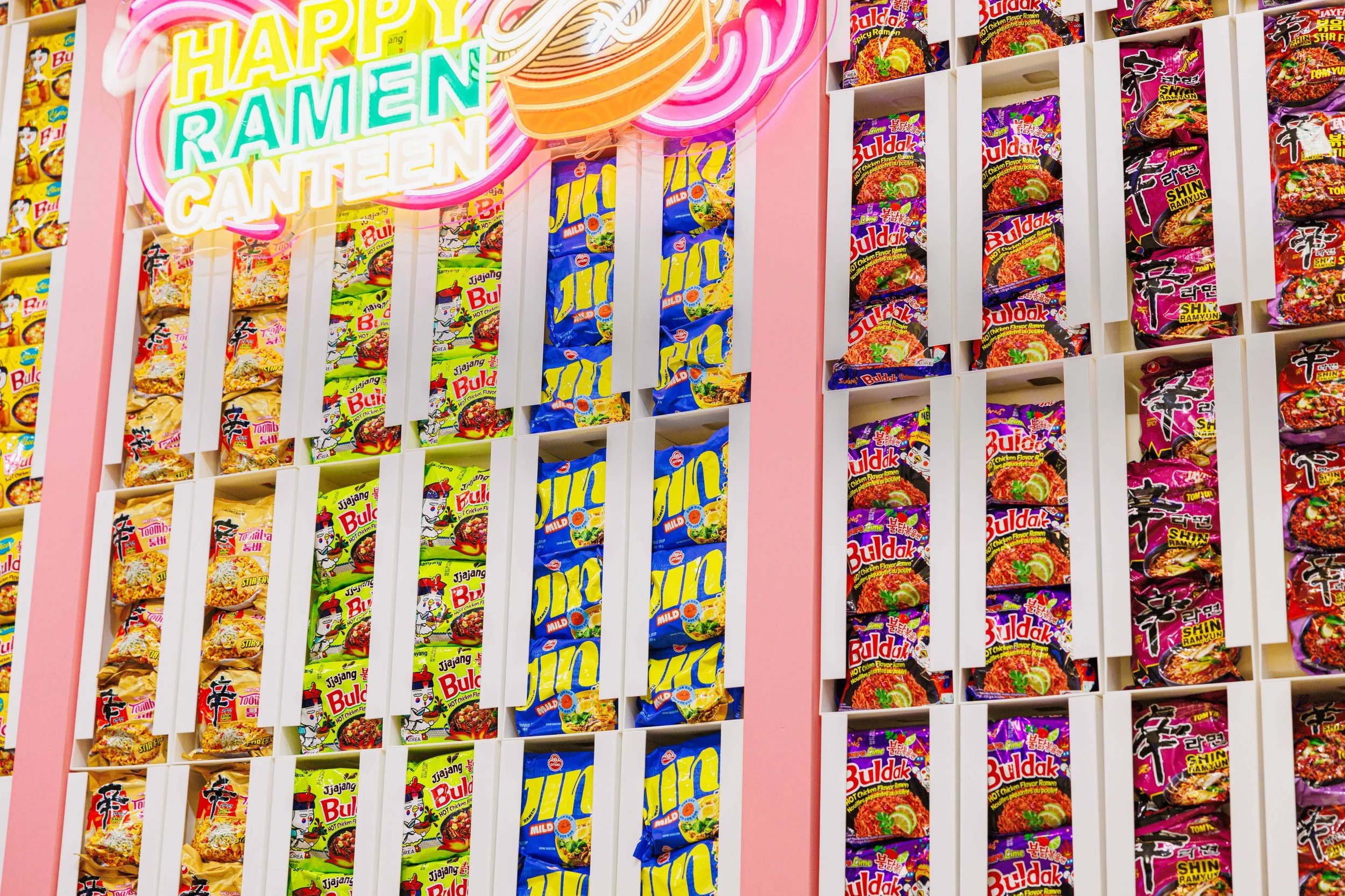 Several shelves stocked with various flavors of instant ramen noodles in colorful packaging at a store. A neon sign reading 'HAPPY RAMEN CANTEN' is above the shelves.