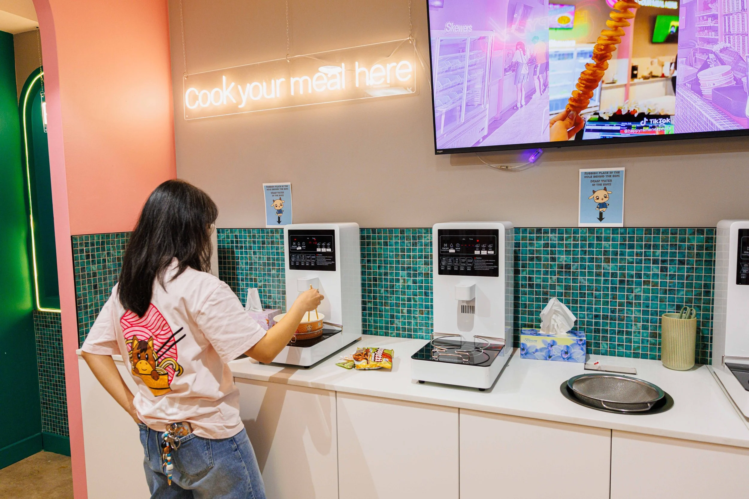 A person standing at a counter in a brightly colored kitchen or food court. The person is wearing a pink t-shirt with a cartoon image on it and denim shorts with a beaded keychain hanging from the pocket. On the counter, there are snacks, a tissue box, and a turntable. Behind the person, there are two paper signs with cartoon characters, a neon sign that reads 'Cook your meal here,' and a large television screen displaying images of a convenience store and a food item.