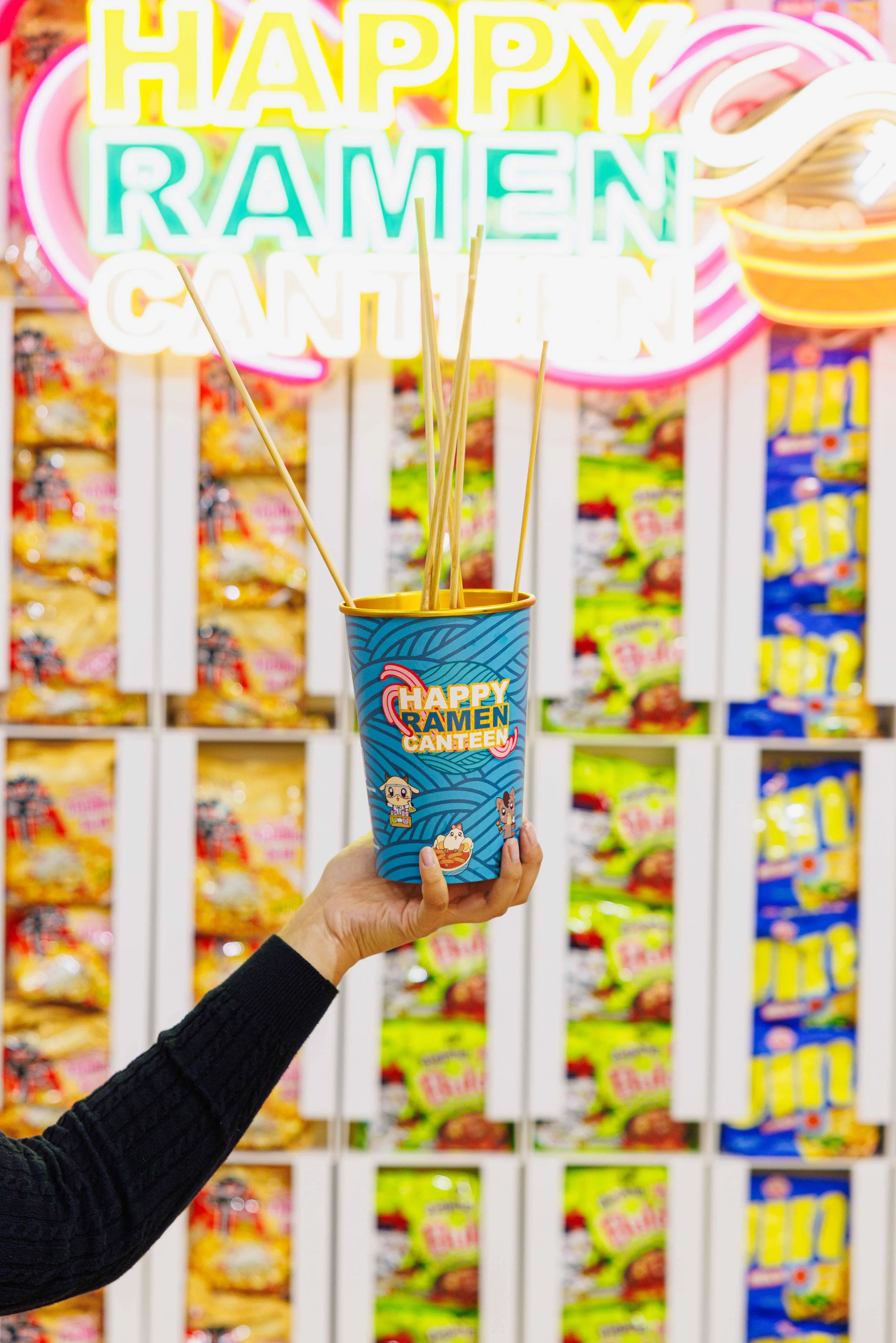 Hand holding a cup with colorful ramen canteen design, with neon sign in the background that reads "HAPPY RAMEN CANTEEN" in a playful, colorful font.