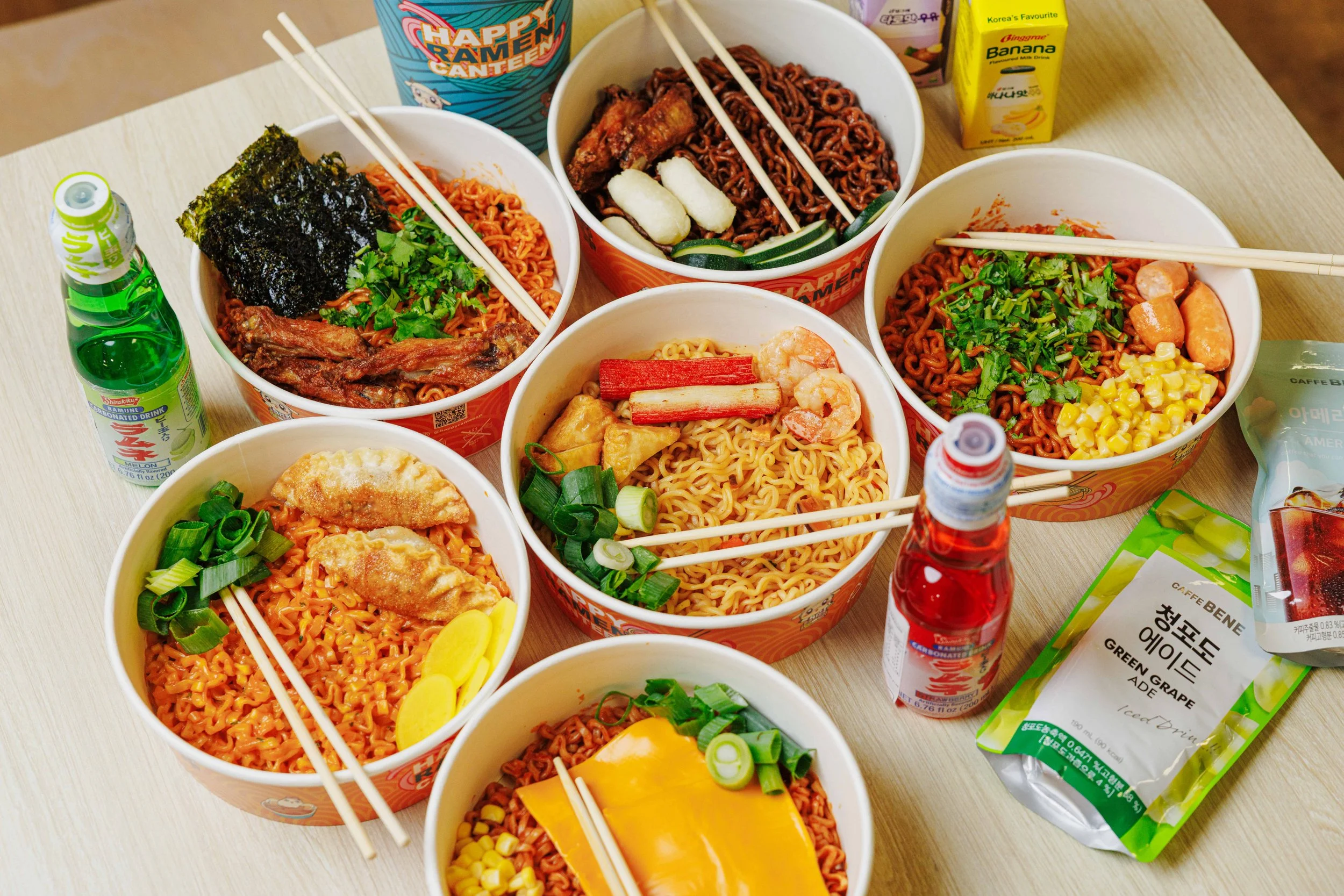 Multiple bowls of ramen noodles with various toppings including seaweed, vegetables, crab sticks, and sausage, on a light-colored table along with drinks and snack packages.