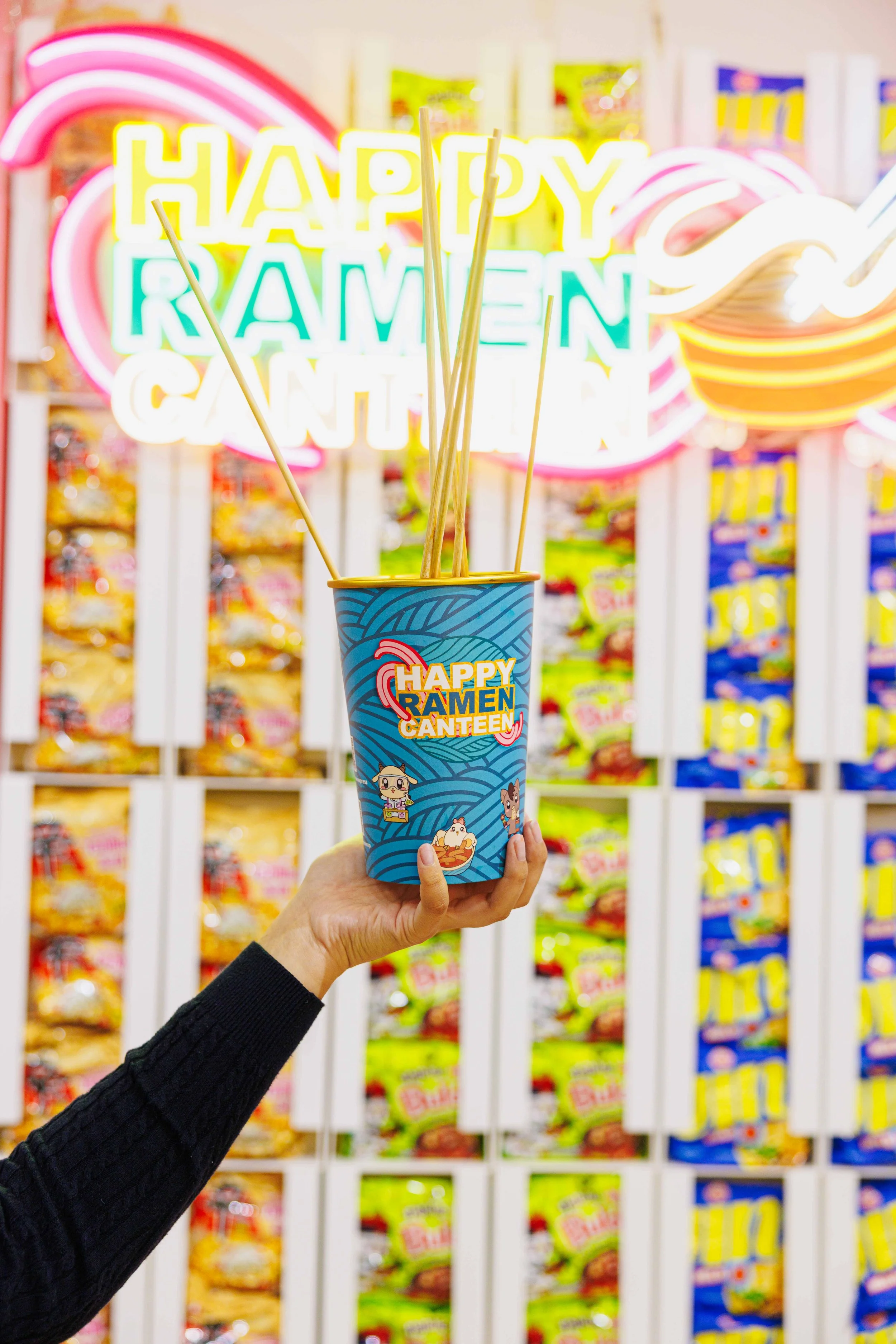 A hand holding a cup with neon signs in the background that read 'Happy Ramen Canteen' and some straws.