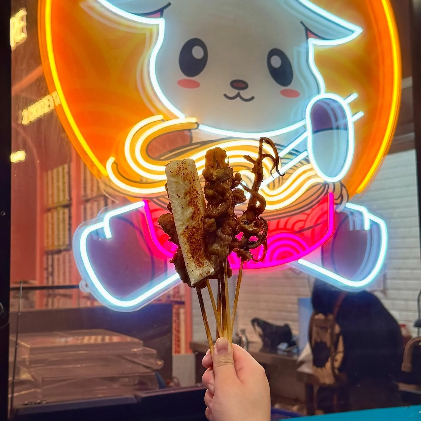 Three skewers of grilled meat and bread held in front of a colorful neon sign featuring a cute cartoon bunny with large eyes, rosy cheeks, and a friendly expression.