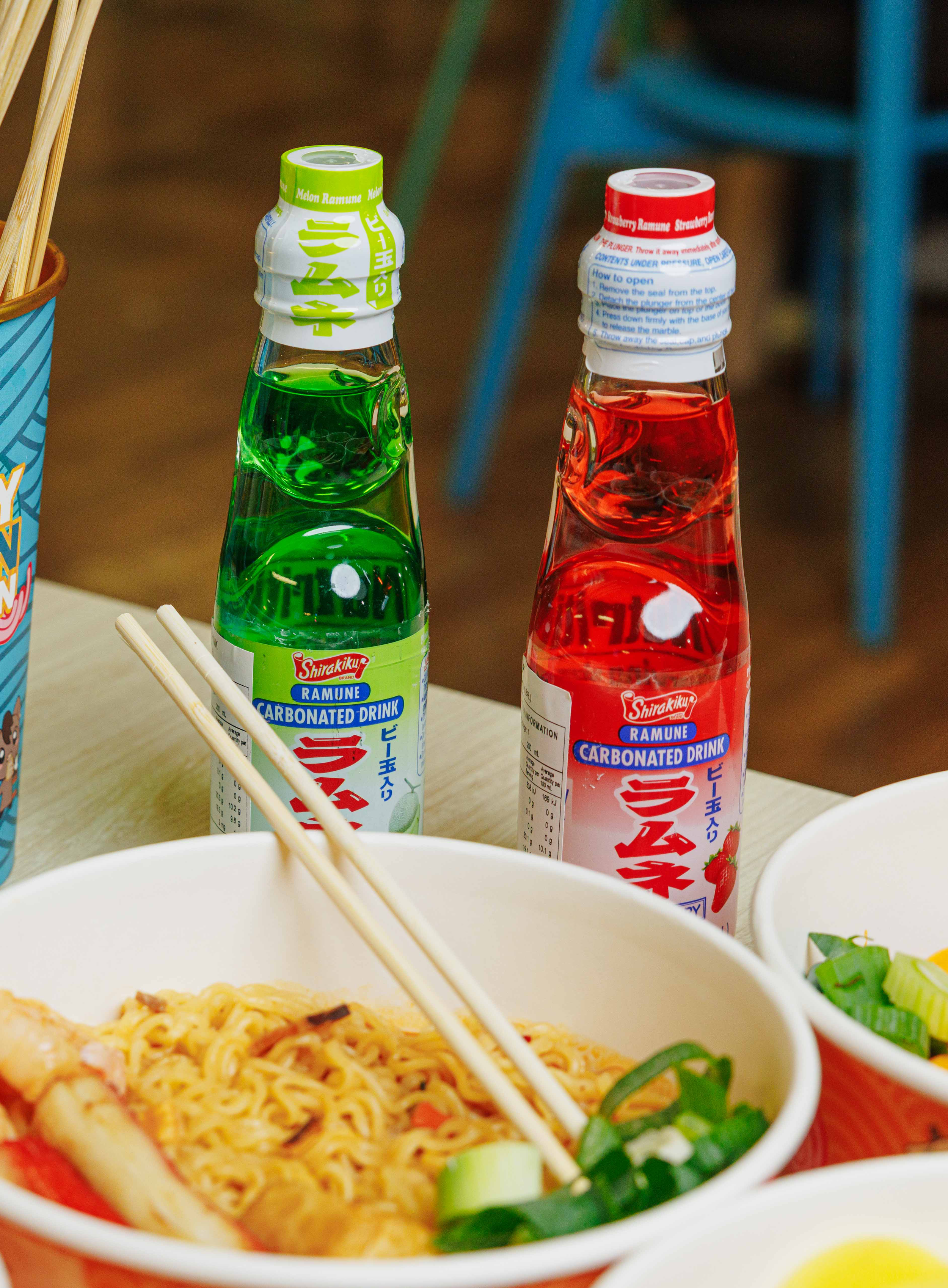 Three bottles of Ramune carbonated drinks, two with green and red labels, on a table with Asian food dishes and chopsticks.