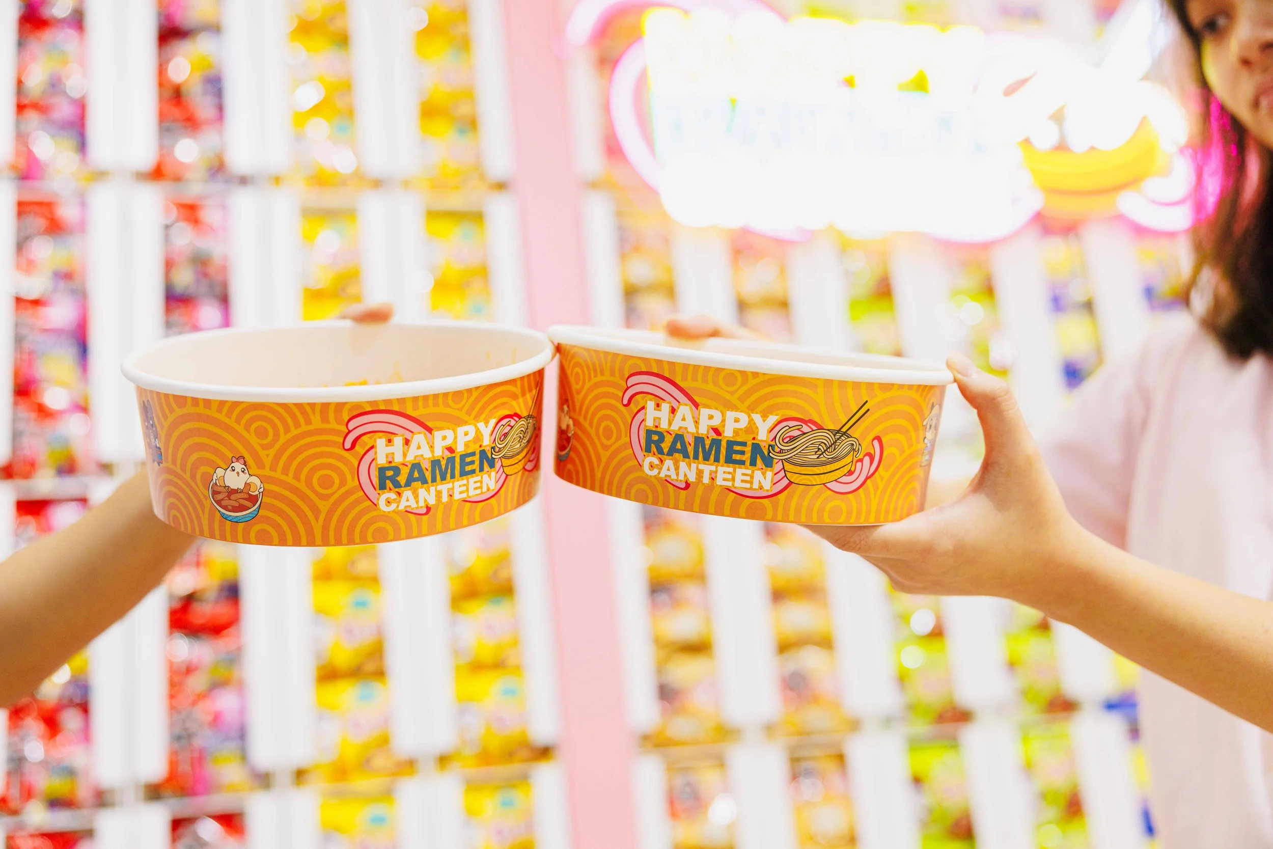 Two people holding cups of ramen at a ramen canteen, with colorful shelves in the background.