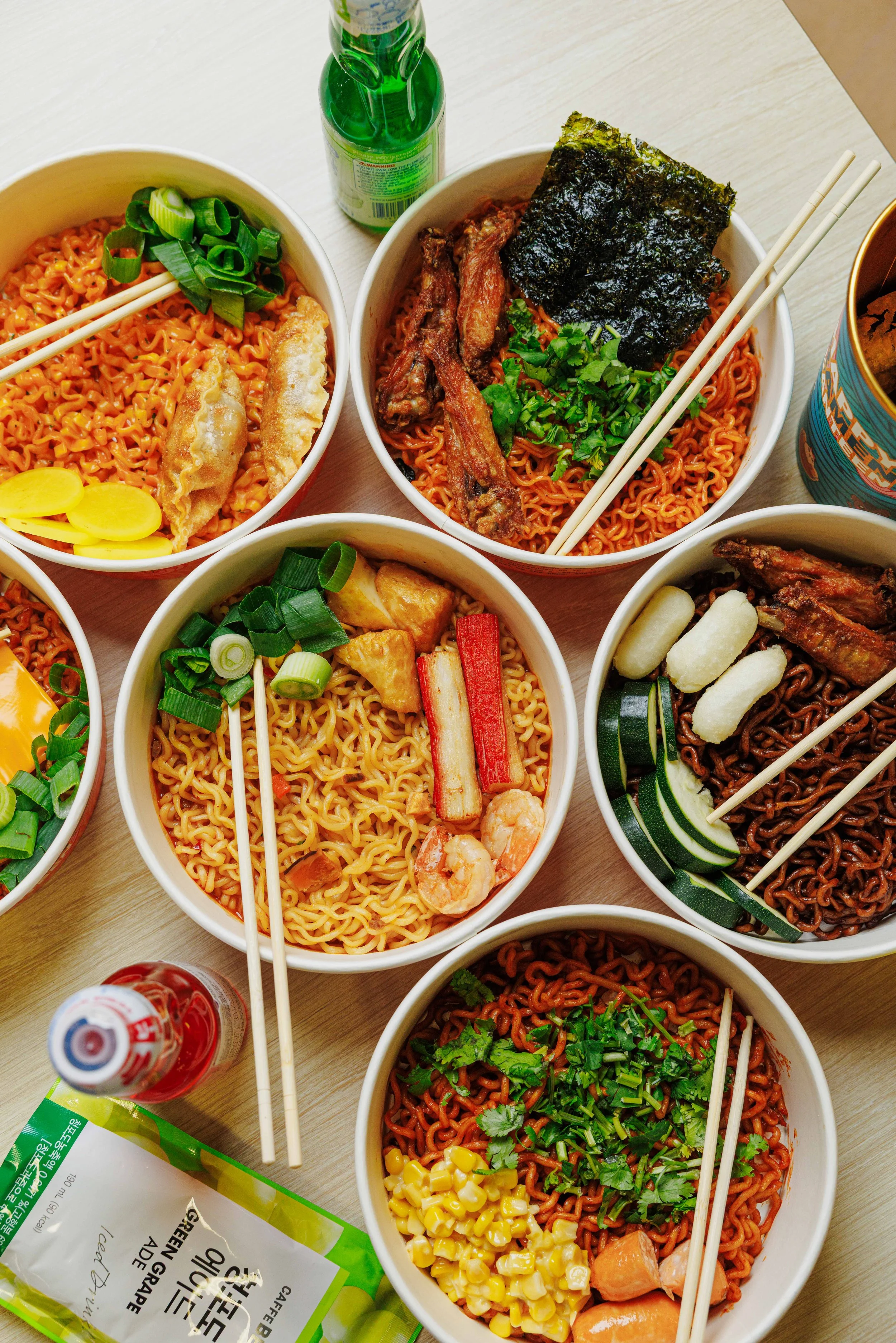 Multiple bowls of different types of ramen with various toppings on a wooden table, alongside a green bottle of soda, a small bottle of red condiment, and a bag of green tea.