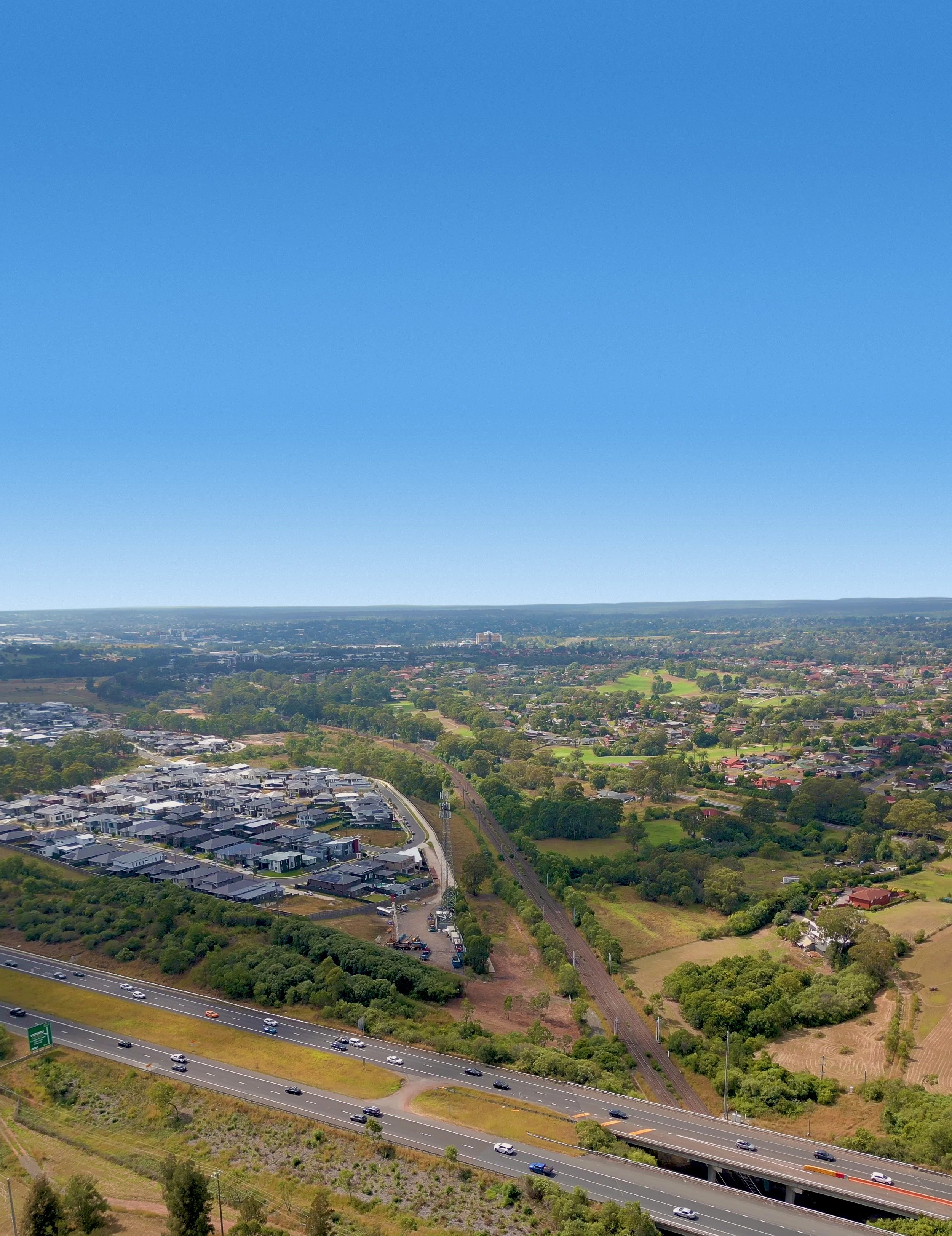 An aerial view of a city with a highway, residential area, and a large amount of greenery, under a clear blue sky.