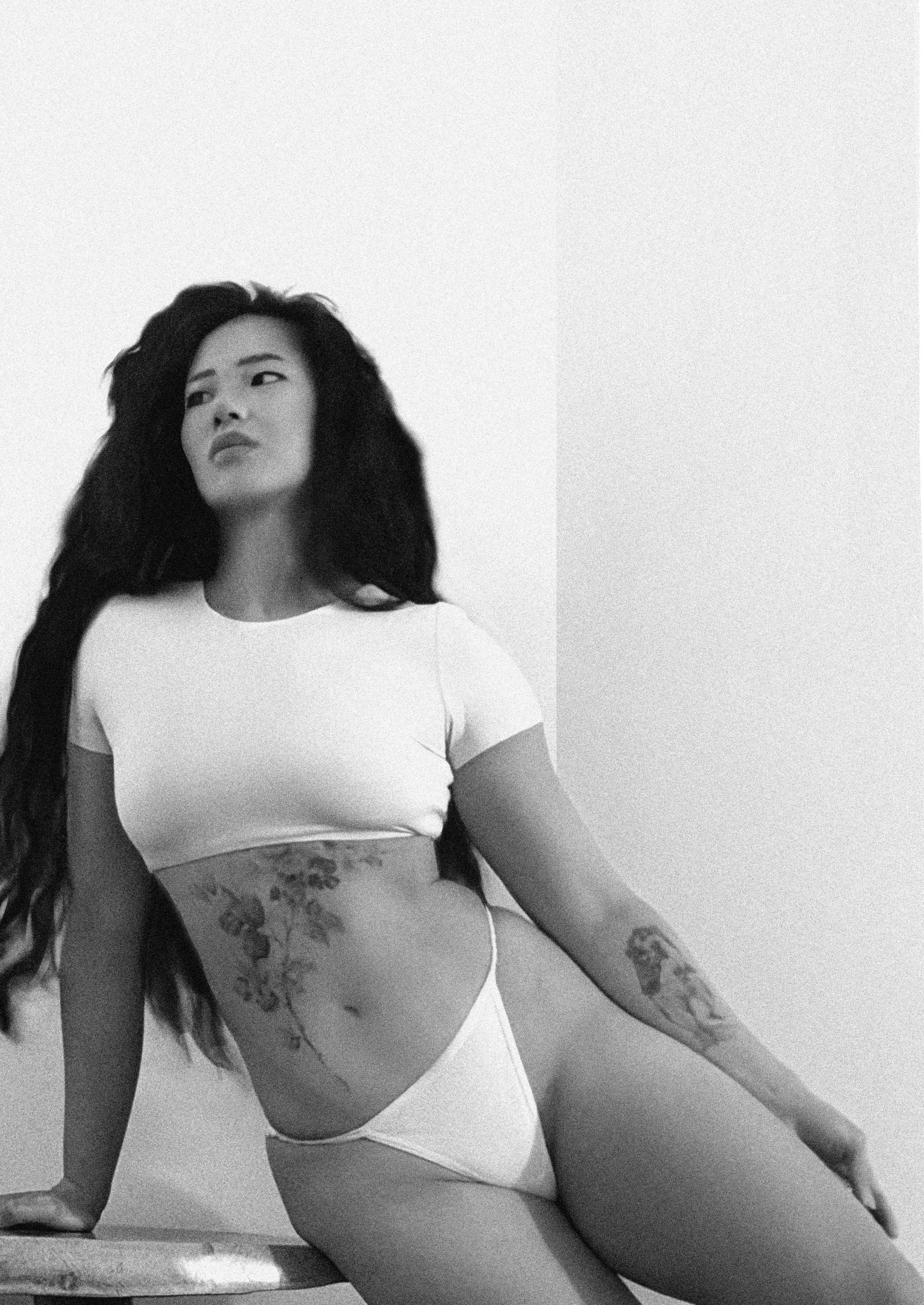 A woman with long dark hair, tattoos on her arms and torso, wearing a white crop top and underwear, sitting on a surface with a plain background.