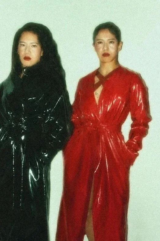 Mizu, NYC dominatrix, standing side by side, both wearing shiny vinyl trench coats, one in black and the other in red, with serious expressions.