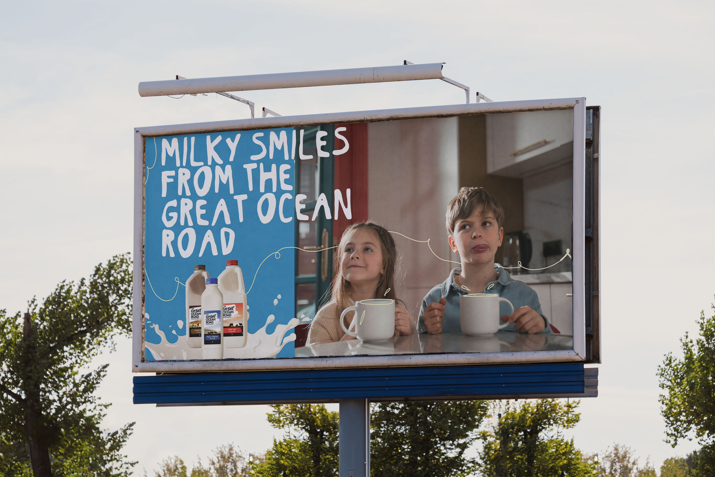 Two children, a girl and a boy, standing behind a milkshake stand with a sign that reads 'Milky Smiles from the Great Ocean Road.' The stand has images of milk cartons on it, and there are trees and a cloudy sky in the background.