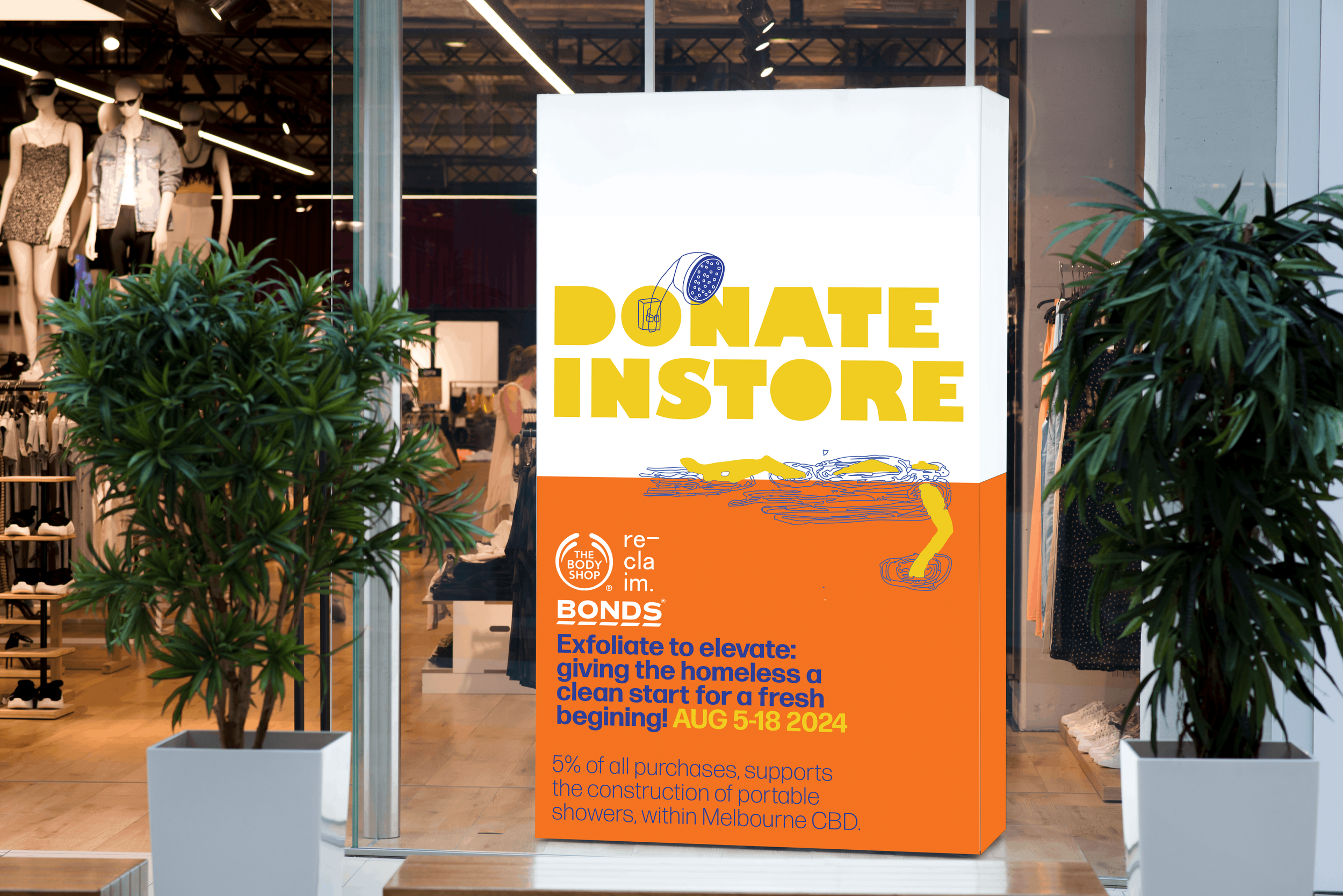 Storefront window with mannequins displaying clothing, green plants on either side, and a large sign promoting a donation campaign from August 5-18, 2024 to support homeless individuals through The Body Shop and Bonds.