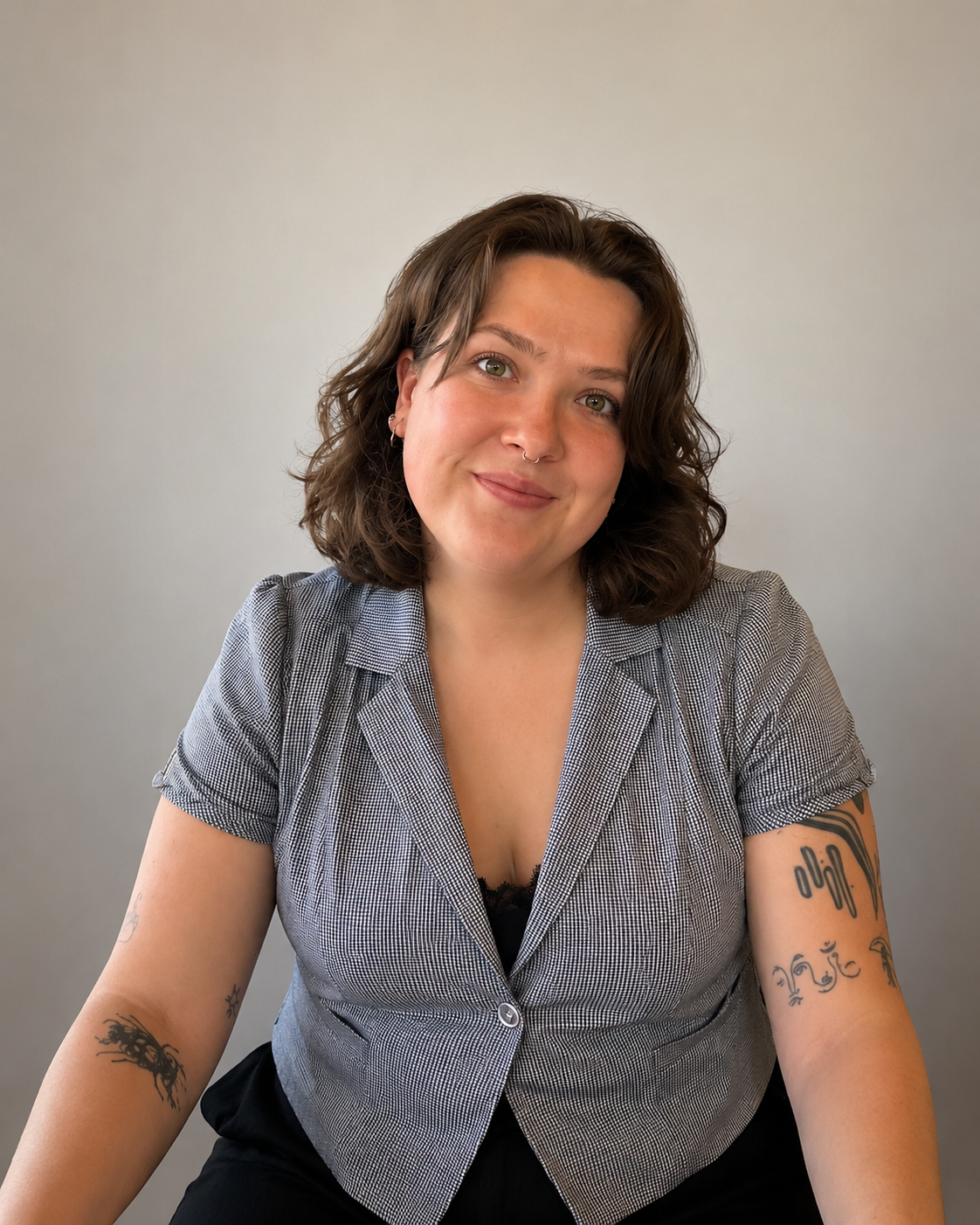 A young woman with shoulder-length curly brown hair, light skin, and tattoos on her arms, sitting against a light gray background. She is smiling softly, wearing a short-sleeved, black-and-white checkered blazer over a black top.